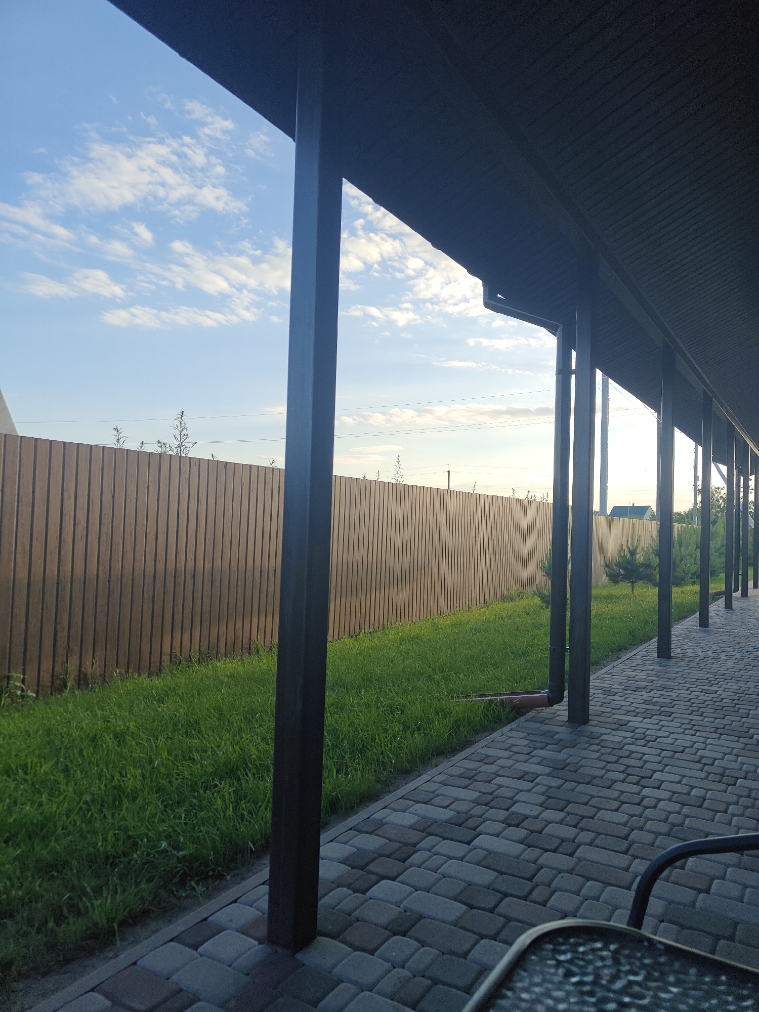 The image depicts an outdoor scene with a covered walkway in the foreground, leading towards the right. The walkway is paved with interlocking gray and brown stones. A dark-colored metal railing is visible at the bottom right. Beyond the walkway, a strip of green grass runs alongside a tall wooden fence. The fence is constructed of vertical planks and stretches horizontally across the frame. Above the fence, a bright blue sky with scattered white clouds is visible. Several dark, vertical posts support the roof of the walkway, which appears to be made of corrugated material. A downspout is attached to one of the posts. Small evergreen trees and some vegetation can be seen beyond the fence. The overall impression is of a residential backyard or a similar outdoor area in a rural or suburban setting. The location is identified as Shkurupiivka, Ukraine. There are no people or active interactions visible in the image.