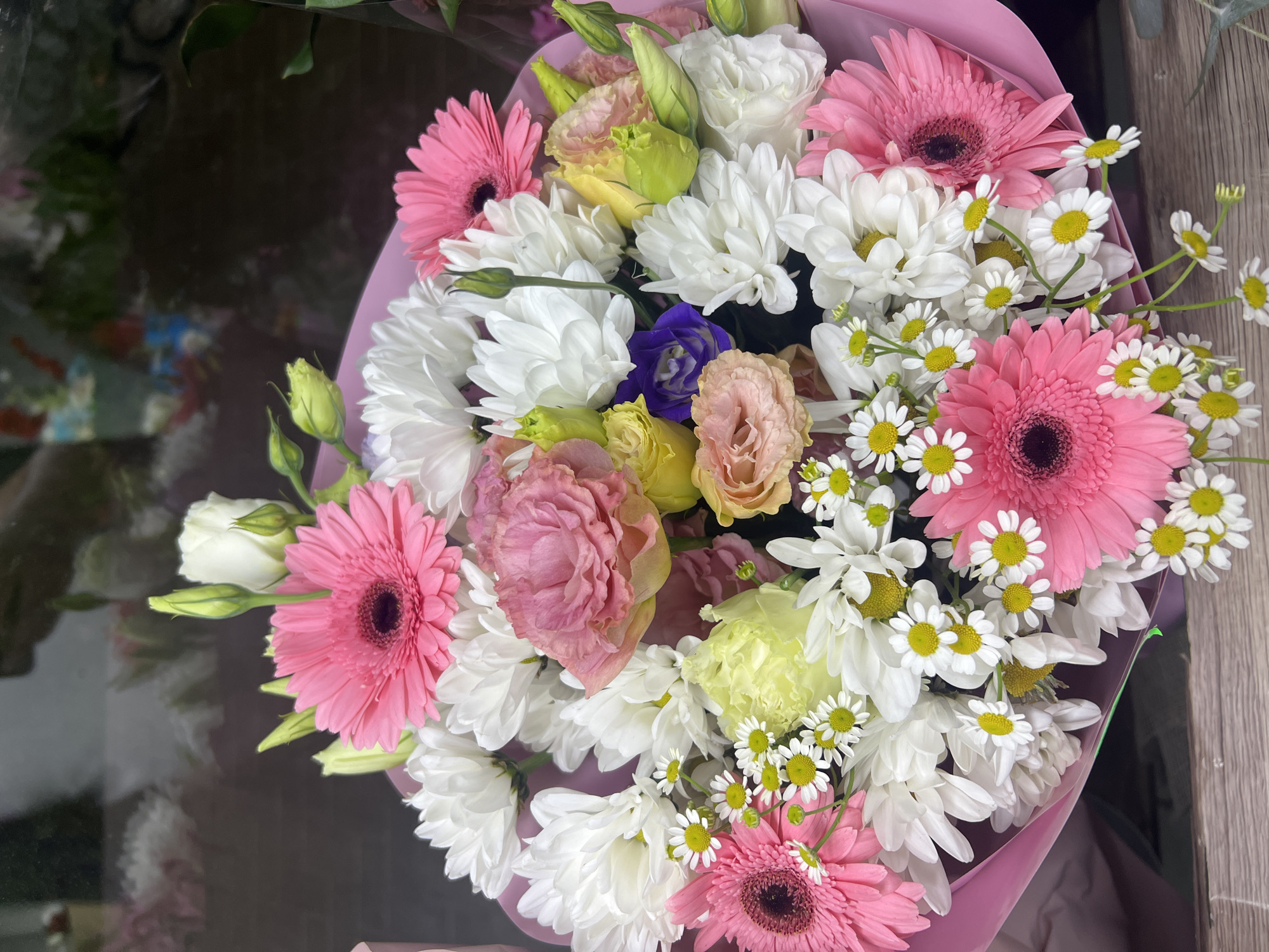 This is a close-up, eye-level shot of a vibrant bouquet of flowers. The bouquet is predominantly pink, white, and yellow, with pops of purple. There are several large pink gerbera daisies with dark centers, interspersed with white chrysanthemums and smaller white daisies with yellow centers. Delicate pink and peachy roses, along with pale yellow and light green lisianthus buds, add further texture and color. The flowers are wrapped in light pink paper, creating a soft, rounded shape. The background is dark and blurred, suggesting an indoor setting, possibly a florist shop or a home. There are reflections in the background, hinting at other objects or the environment outside of the frame. The lighting is bright, illuminating the details of the flowers. The scene appears to be staged for a photograph, showcasing the beauty and arrangement of the floral display. There is no indication of the specific location within Chișinău, Moldova.