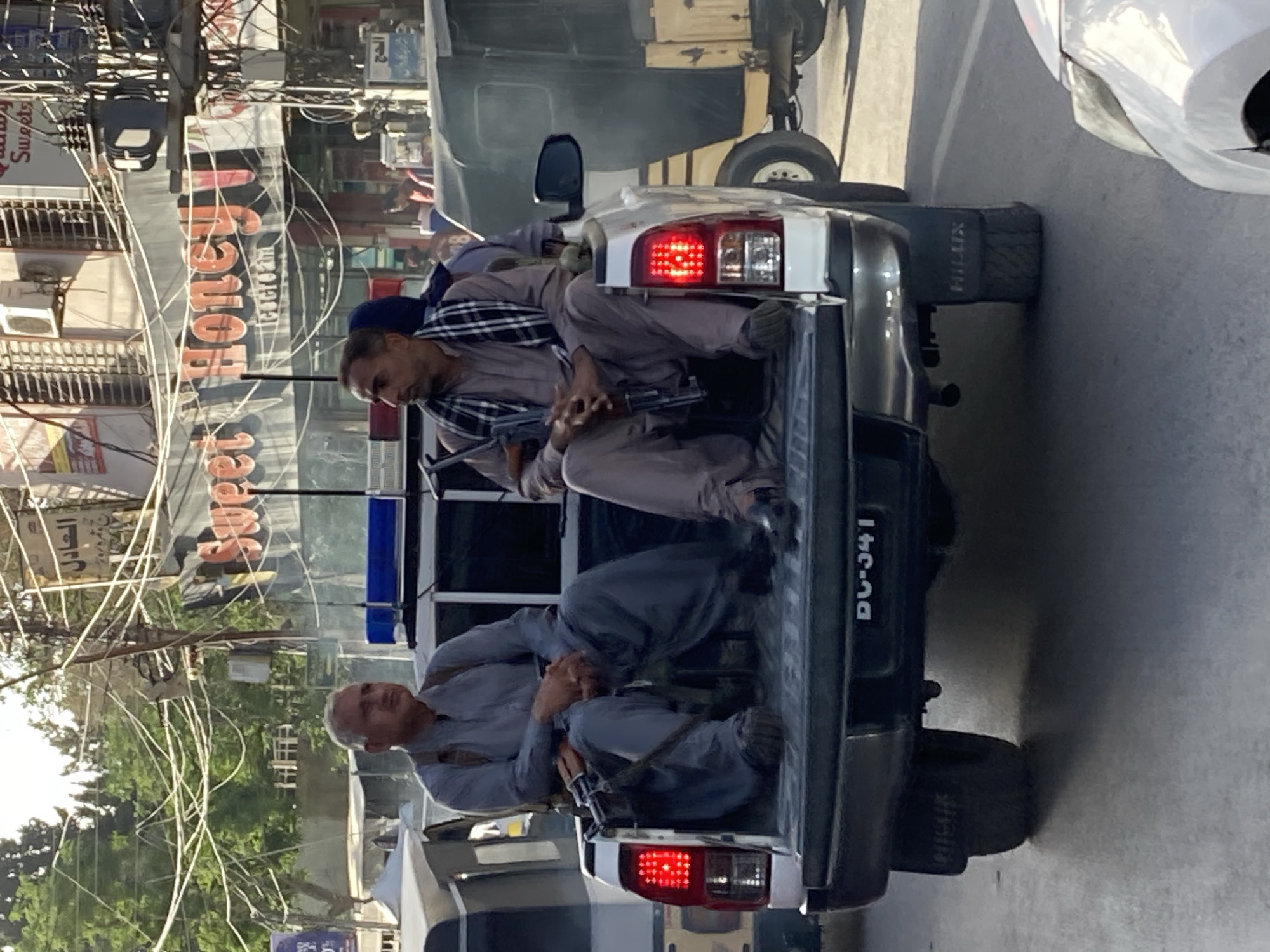 Two armed men sit in the back of a pickup truck, one wearing a blue patterned scarf and the other suspenders. The truck has flashing blue lights on the cab and a license plate visible on the rear. The scene appears to be set in an urban environment, with buildings and overhead wires visible in the background. A sign with the word "Honey" in English can be seen in the background. The location is Quetta City Tehsil, Pakistan.