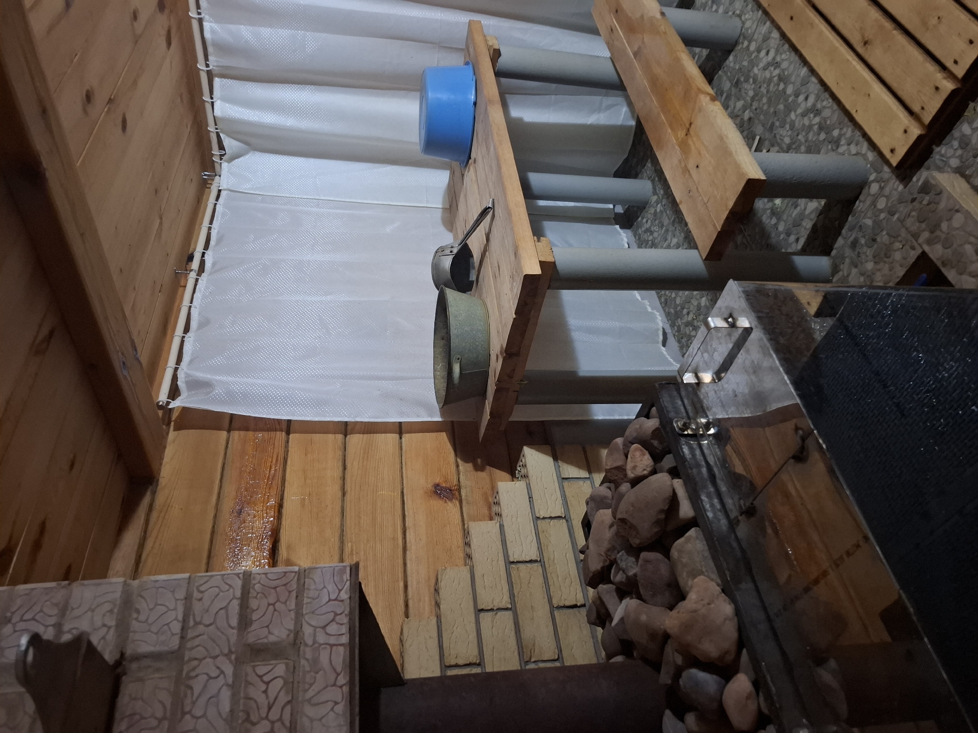 This indoor shot depicts the interior of what appears to be a sauna or steam room in Амзя, Russia. The walls are primarily constructed of light-colored wood paneling. A white, translucent curtain-like material hangs along one wall.
There's a wooden structure that serves as a shelf or support, upon which a blue plastic bucket and metal scoop are placed. The structure also incorporates gray cylindrical supports. The flooring consists of various materials, including patterned tiles near the foreground, transitioning to wooden planks, then bricks leading to a stone hearth or rock-filled enclosure. A dark-colored metal object, likely part of the sauna's heating system, is positioned near the rocks.