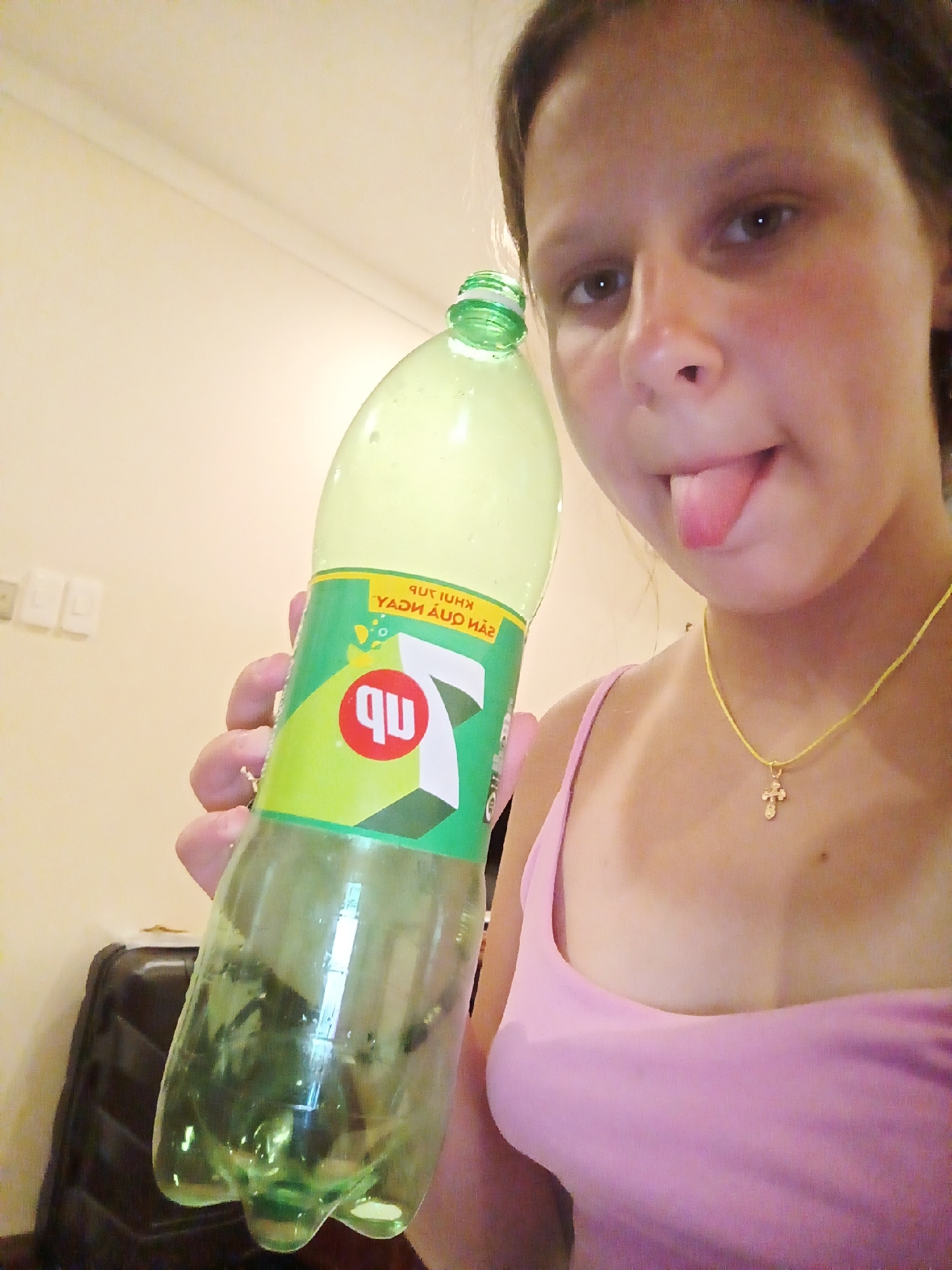 The media file shows a young person holding a green plastic bottle of 7UP. The person is facing the camera and sticking their tongue out playfully. They are wearing a pink tank top and a thin yellow necklace with a small cross pendant. The background is a plain, light-colored wall. The lighting suggests it might be daytime or indoors with artificial light. The setting appears to be a home or indoor environment. Text visible on the bottle includes "7UP" and what appears to be Vietnamese text: "7UP KhUI TuH" and "YADU AUQ HAU". The overall mood is lighthearted and casual.