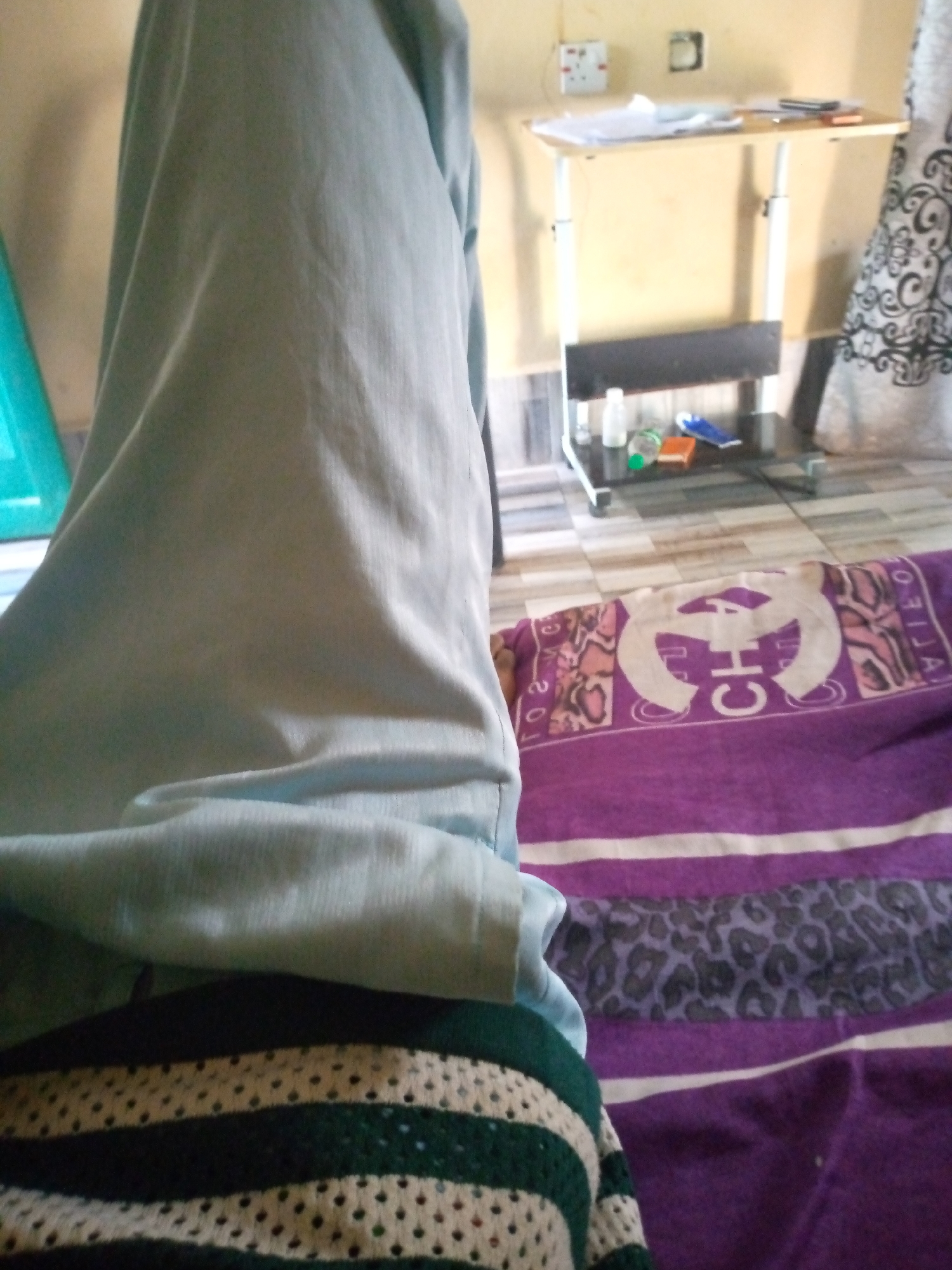 The image depicts an indoor scene. The foreground is dominated by a person's legs covered in light blue fabric, partially obscuring a bed with a purple and white striped blanket. A dark green and beige striped fabric is visible in the lower left corner. In the background, a portable height-adjustable desk with a light-colored wooden top is positioned against a light-colored wall. Papers and a mobile phone are on the desk. Below the desk, a dark shelf holds a clear plastic bottle, an orange object, and a blue object. A patterned curtain is visible on the right side of the image. The floor is tiled in a checkered pattern. The setting appears to be a room in Jalingo, Nigeria.