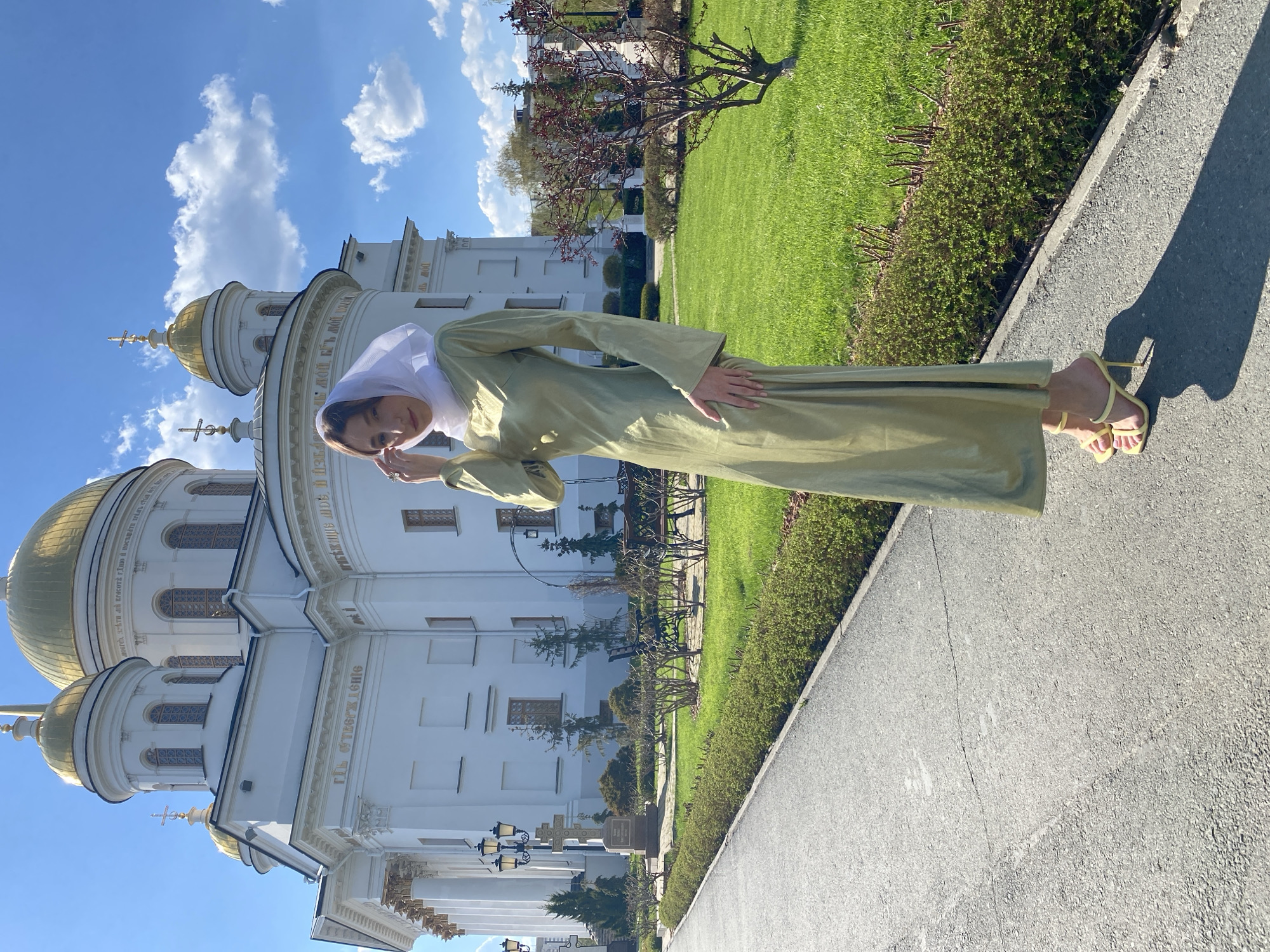 A woman stands on a sidewalk adjacent to a green lawn, posing in front of the Church on Blood in Yekaterinburg, Russia. She is wearing a light green, long-sleeved dress and a white head covering, complemented by yellow sandals. The church, a prominent white structure with golden domes, stands against a bright blue sky with scattered clouds. A pathway separates the sidewalk from the lawn, which is lined with bushes and trees.