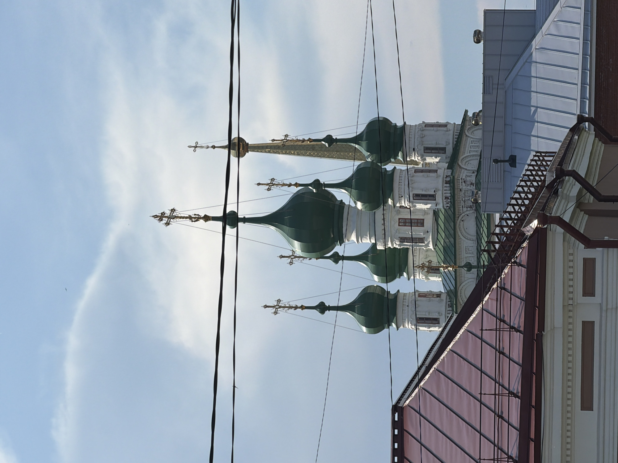 The image captures a view of the Holy Trinity Cathedral in Solikamsk, Russia, as seen from behind some foreground buildings and power lines. The cathedral features multiple green, onion-shaped domes topped with golden crosses, and one prominent golden spire. In front of the cathedral, there are rooftops of various buildings, one with a reddish-brown metal roof and another with a grey metal roof. The sky is a pale blue with scattered white clouds. The scene appears to be taken during the daytime under clear weather conditions. There are no people visible in the image.