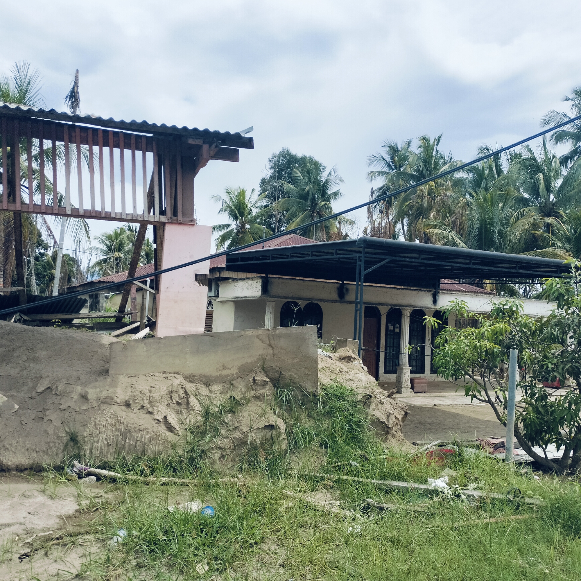 This image depicts a scene of tropical residential buildings amidst significant ground disturbance, suggesting the aftermath of a natural event in Manyang Cut, Indonesia. The setting is daytime, under a light blue sky with scattered white clouds, indicating an overcast or partly cloudy day.
In the immediate foreground, there are mounds of textured, light brown dirt or sand, showing signs of erosion and displacement. Green grass and weeds are growing through these mounds and across the uneven ground, which appears somewhat muddy. Scattered debris, including pieces of wood and plastic bottles, lies on the ground.
Dominating the left side of the mid-ground is a damaged wooden structure, possibly part of a gate, fence, or an elevated walkway, topped with a corrugated metal roof section. Its wooden planks are vertically oriented, and some appear broken or askew. Behind and to the left of this structure, another house with a reddish-brown corrugated roof is partially visible, obscured by palm trees.
On the right side of the mid-ground and extending into the background is a house with a light-colored, possibly stucco or concrete, facade. It features dark, arched window or door openings and decorative white columns supporting a small porch area. A dark, modern-looking metal car-port or awning structure extends over the front of this house. Electrical wires are strung across the middle of the frame, passing above the houses and through the trees.
The background is filled with numerous palm trees, characteristic of a tropical environment. Some appear lush and green, while others, particularly towards the right, show signs of damage or dryness. A small, leafy tree, possibly a mango tree, stands in front of the house on the right. No people or visible text are present in the image. The overall impression is one of disruption and recovery following a significant environmental event.