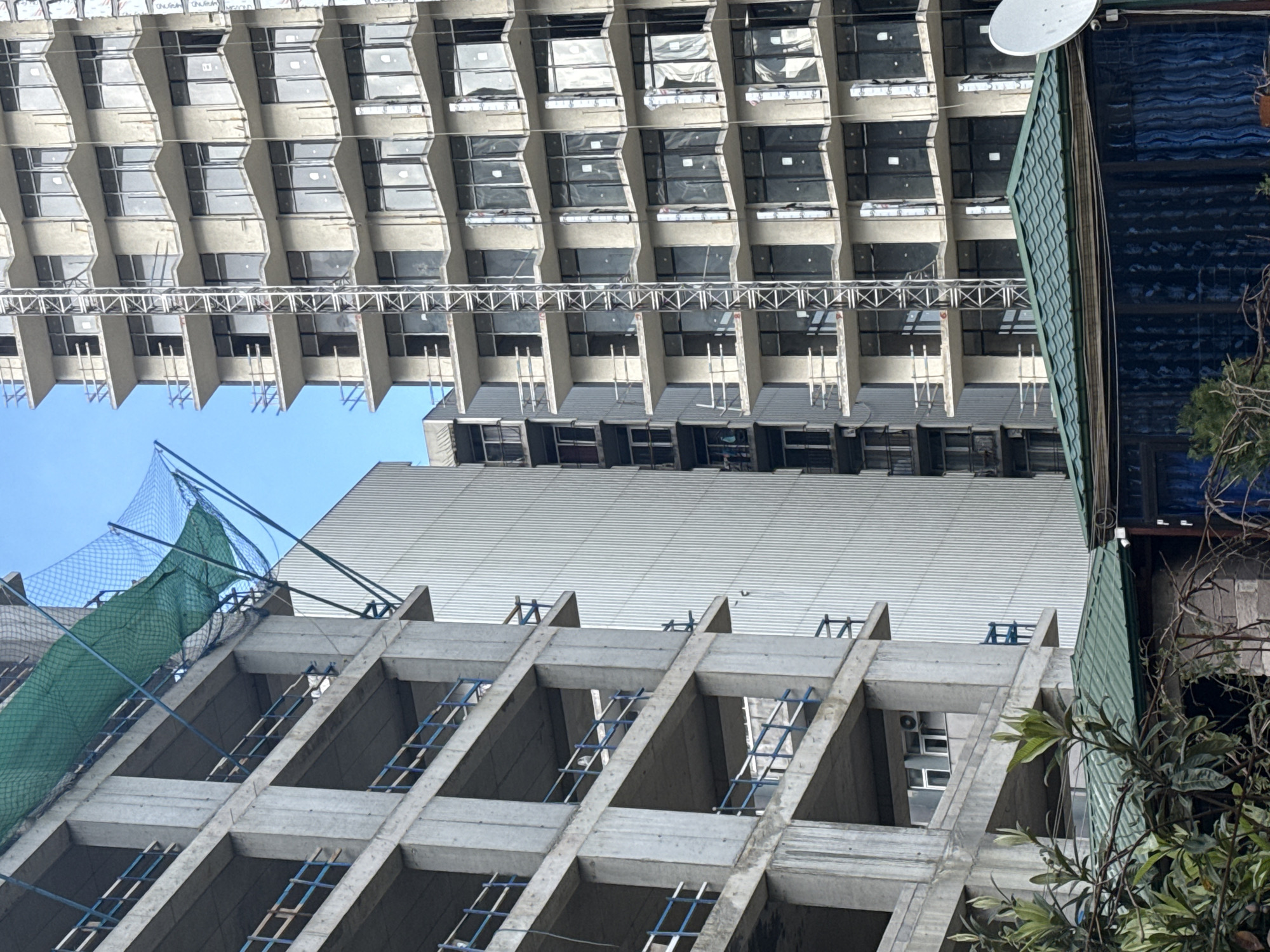 The image shows two buildings in Batumi, Georgia. The building at the bottom of the frame is under construction. It is a concrete structure with rebar visible. A green safety net is draped on part of the building's facade. The other building is completed and is in the background. It is a tall, modern building with a repetitive pattern of windows and balconies. A satellite dish is attached to the roof of the second building on the right. In the middle of the two buildings is a corrugated metal roof. Some trees and plants are on the right side of the frame. The sky is blue and clear.