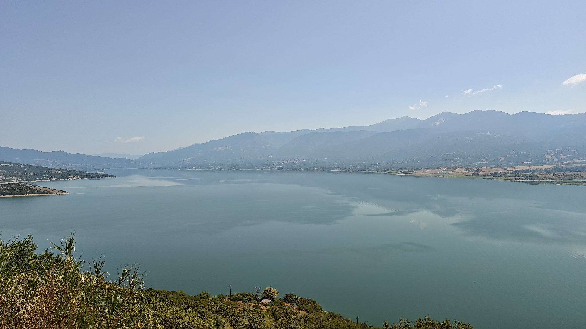 An expansive, tranquil lake dominates the scene, its surface reflecting the clear, pale blue sky and the distant, hazy mountain range. The mountains, layered in varying shades of blue and green, stretch across the horizon, their peaks softened by atmospheric perspective. Along the far shore, the land appears arid and populated with scattered vegetation and some signs of human settlement, like faint structures and roads visible in the distance. The foreground is occupied by lush, green vegetation and some dry, yellowed grasses, suggesting an elevated viewpoint overlooking the lake. The water itself is a deep, serene blue-green, with subtle ripples and the faint impression of clouds on its surface. A small, rocky peninsula juts out into the lake on the left side of the image. The overall impression is one of natural beauty and peaceful isolation. The image depicts the artificial lake of Polyfytos, a reservoir on the Aliakmonas River located within the Servia Municipal Unit, Greece.