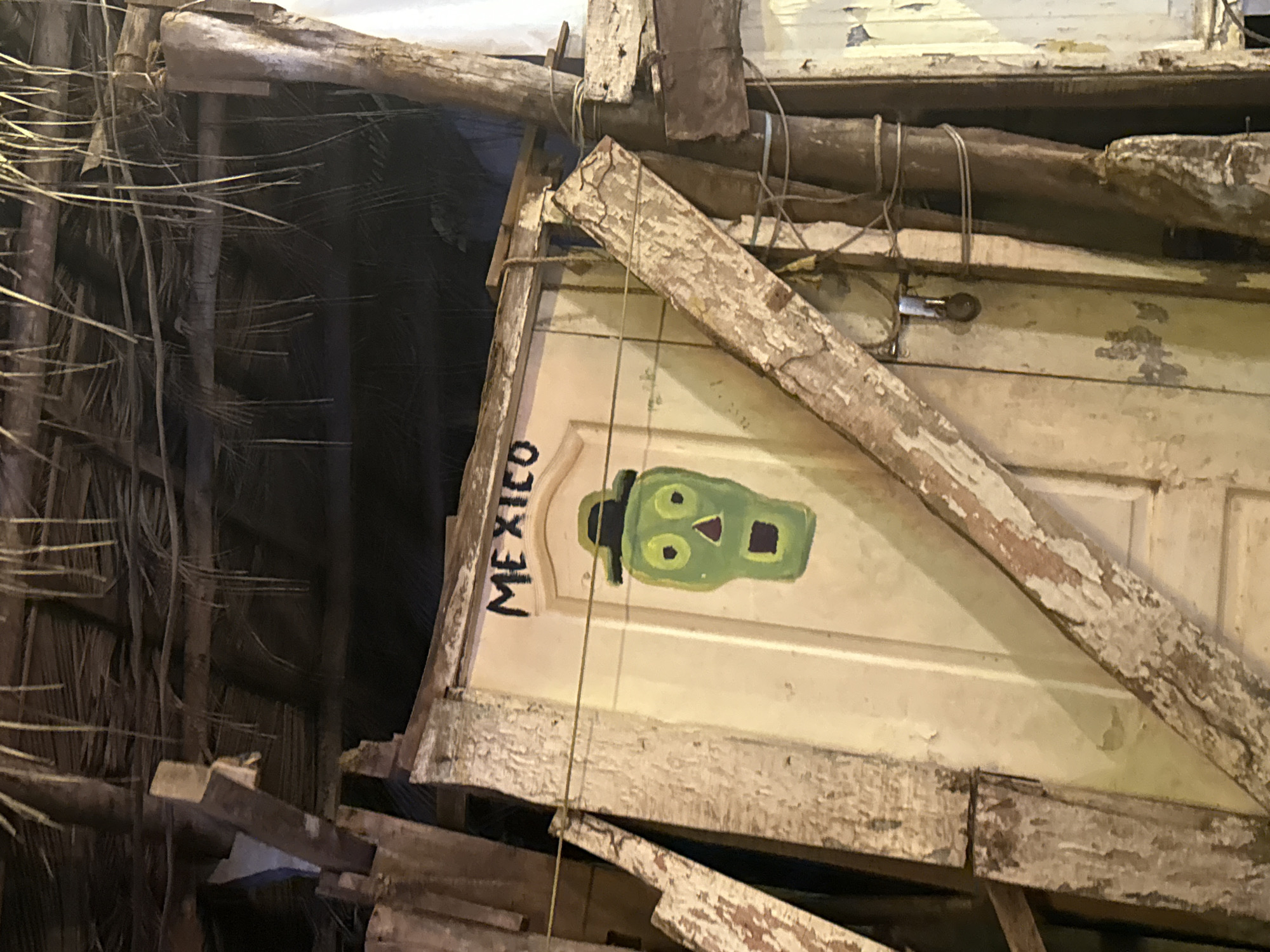 Image shows part of a structure, possibly a building or shelter, made from reclaimed and weathered materials in what could be a makeshift or rustic setting. The structure is constructed from wooden planks, beams, and possibly recycled doors, all showing signs of age and wear. The word "MEXICO" is written vertically in black on the door, along with a crude painting of a green human-like head.

The presence of thatching or similar natural material on the left side of the frame suggests a tropical or rural environment, with the overall ambiance being somewhat rundown or neglected. The lighting seems dim, indicating either nighttime or an indoor space with poor illumination. The setting is cluttered and appears to be in the vicinity of Chapora, India.