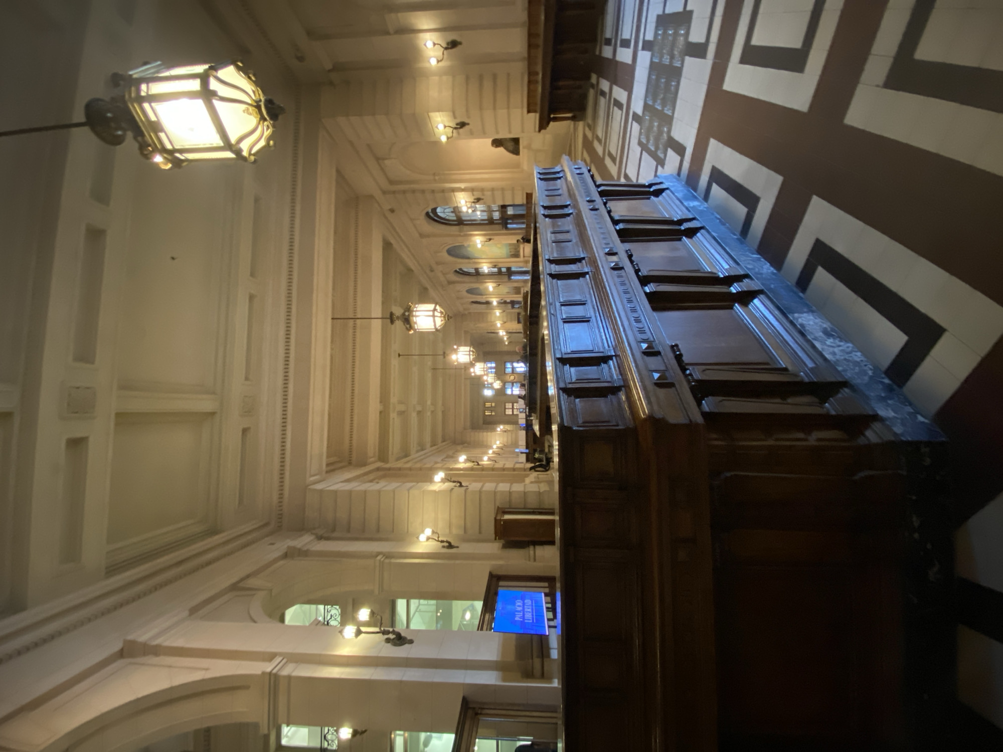 The image shows the interior of the Palacio de la Legislatura de la Ciudad de Buenos Aires in Buenos Aires, Argentina. The scene is an elongated hallway with a high, ornately decorated ceiling and walls featuring arched windows. A row of antique-style lanterns hangs from the ceiling along the hallway. Along one side of the hallway runs a long, dark wooden desk or counter. The floor features a geometric pattern of dark and light tiles. A screen displays the words "Palacio Libertad".