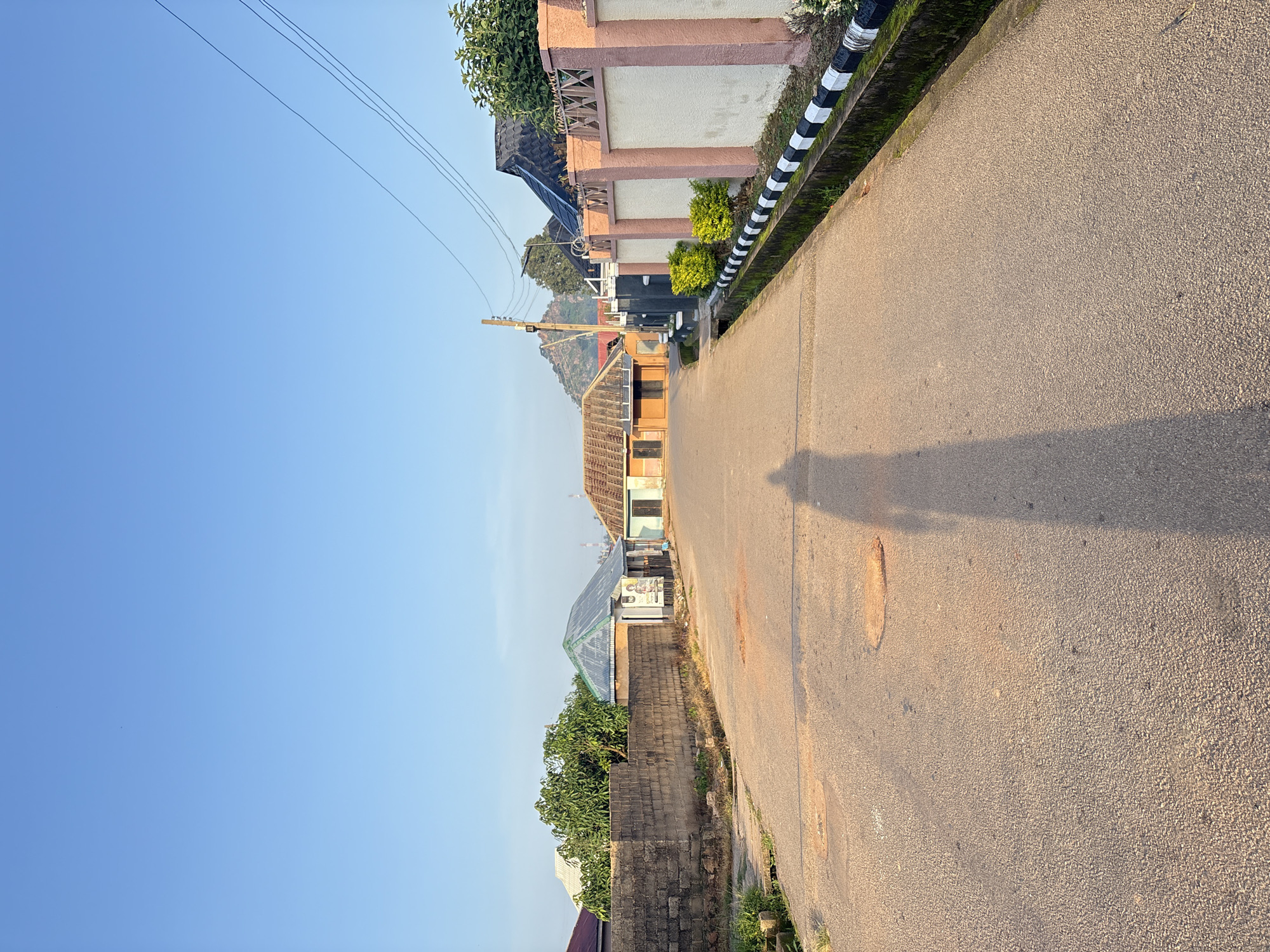 The image depicts a residential street in Bukuru, Nigeria, under a clear blue sky. The road, paved with asphalt, shows signs of wear with several circular patches of exposed earth. The right side of the road is bordered by a decorative wall with a pale beige and pink color scheme, topped with a textured pattern. Small green bushes are planted along the base of this wall. On the left side of the road, a lower brick wall separates it from properties with buildings behind. Several houses are visible, varying in architectural style and roofing materials, including corrugated metal and tiled roofs. Trees, including a large one with lush green foliage, are present. Electrical wires stretch across the sky, supported by a utility pole. The shadow of a person, likely the photographer, is prominently cast on the road, stretching from the foreground towards the middle of the image. The overall scene suggests a quiet, daytime environment in a residential area.