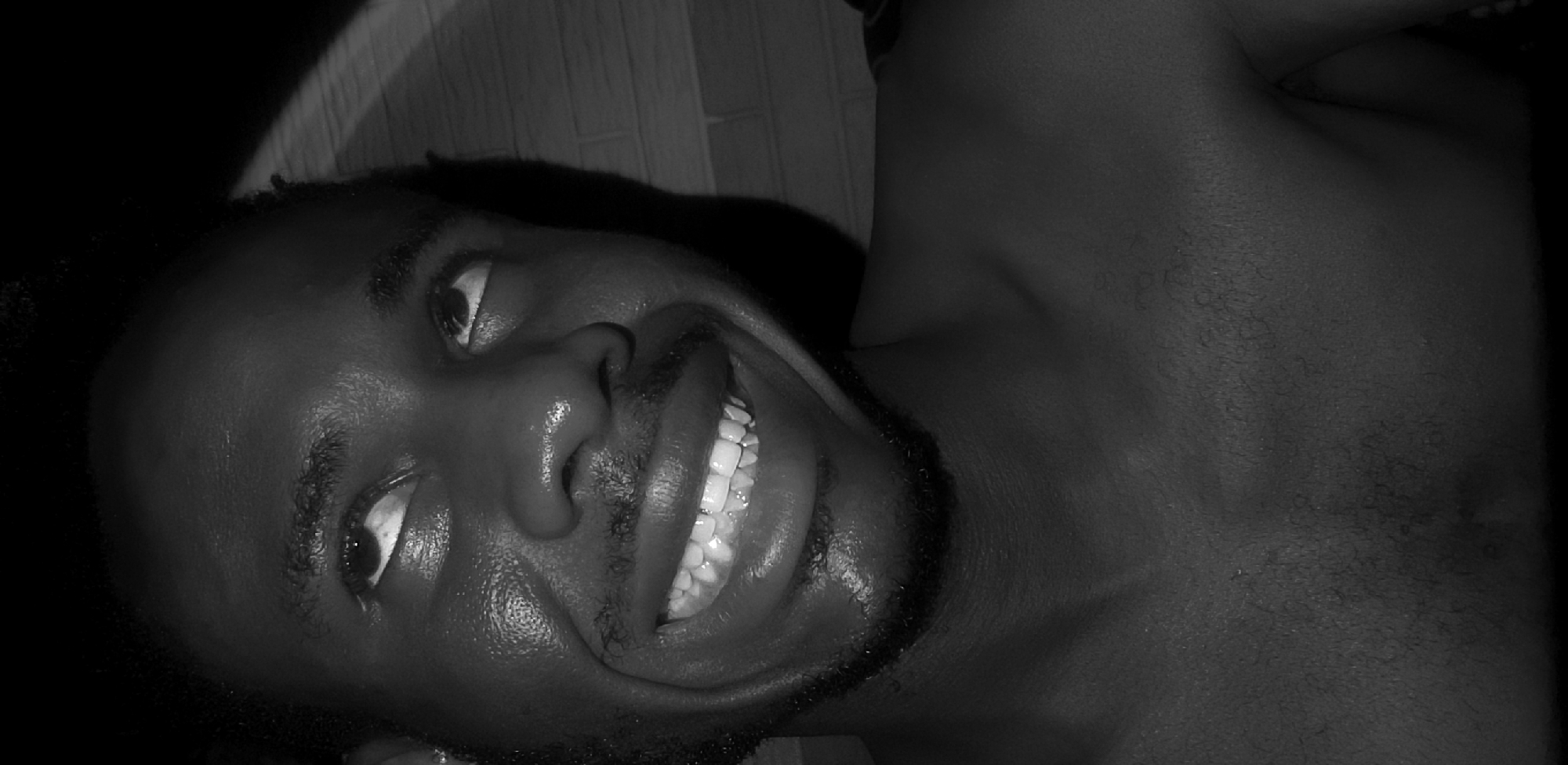 A black and white, close-up portrait features a young adult, likely male, with dark skin, lying down and looking upwards with a broad, joyful smile. The subject’s face is angled towards the top right, dominating the left side of the frame. His eyes are wide open, gazing to the side, and his mouth is open in a wide grin, revealing numerous bright white teeth. He has short, dark, curly hair and a neatly trimmed beard. The skin on his forehead, nose, and lips exhibits a noticeable sheen, suggesting strong, direct lighting or perspiration.
The photograph is captured from a low angle, accentuating the subject's relaxed posture. His bare shoulder and upper chest are visible on the right side of the frame, indicating he is shirtless or wearing minimal attire. The background is dark and out of focus, with a faint textured pattern discernible behind his head, possibly a wall or floor. The black and white rendering creates high contrast, with bright highlights on the face juxtaposed against deep shadows, particularly behind the head and on the left side of the face. The expression conveys a sense of happiness and cheerfulness. No text is visible within the image. The location context provided is Narayi, Nigeria.