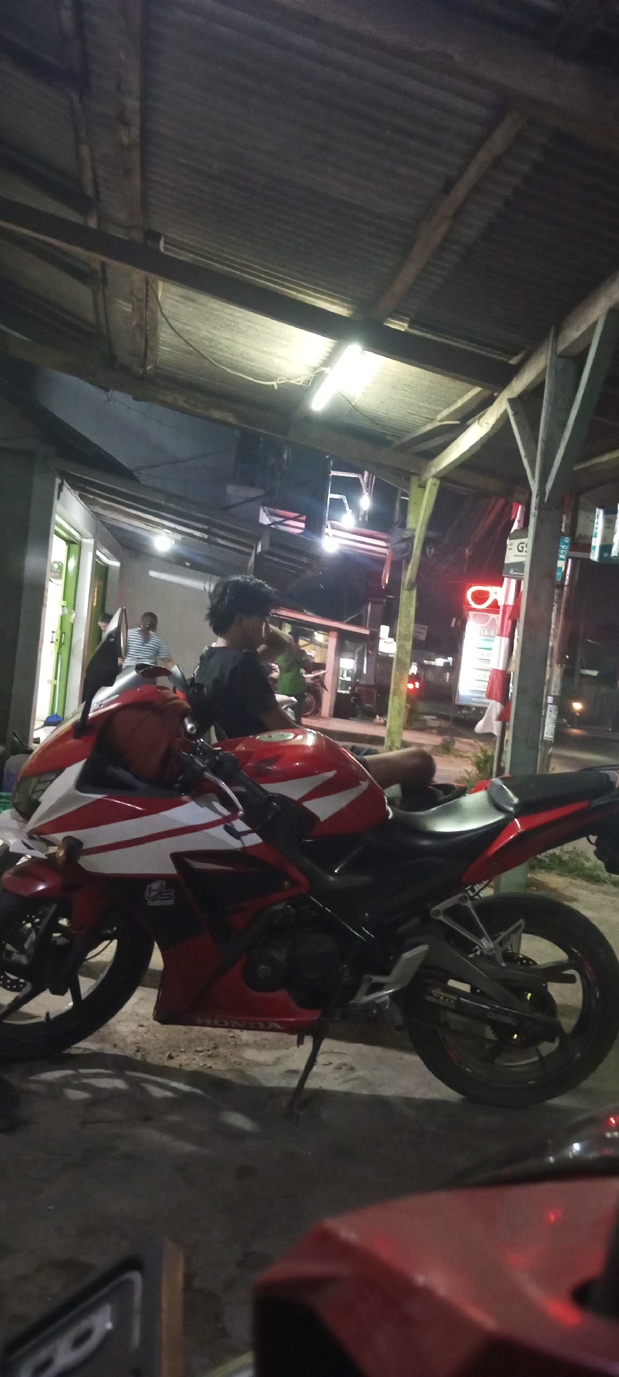 A red and white motorcycle is parked on a concrete surface under a corrugated metal roof supported by wooden beams. The motorcycle is in the foreground, with its front wheel on the left and rear wheel on the right. A person with dark, curly hair is sitting on the motorcycle, facing away from the camera. In the background, several other people are visible, some standing and some sitting. There are also buildings with open doorways, fluorescent lights, and a sign that appears to be in Indonesian. The scene suggests a street-side or roadside setting, possibly in a market or a commercial area in Setu, Indonesia.