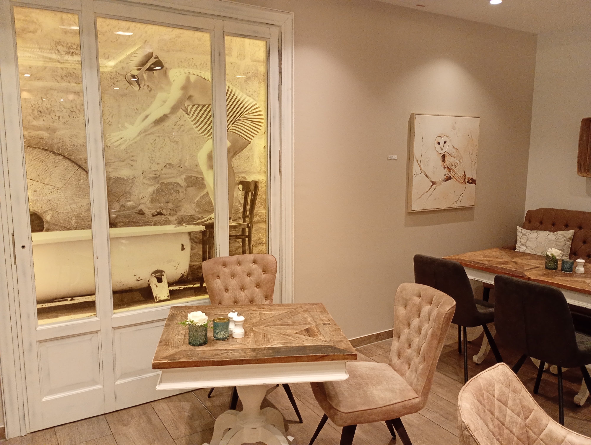 The image depicts the interior of a cafe, likely in Bad Sachsa, Germany, as indicated by the presence of decorative elements suggestive of a spa or wellness theme. A large glass-paneled door, painted white, dominates the left side of the frame. Behind the glass, a vintage clawfoot bathtub is visible, accompanied by a large, textured stone wall and a vintage millstone. A monochrome mural of a person in a striped swimsuit and swimming cap, posed as if diving, is superimposed on the scene behind the glass.
In the foreground, a rustic wooden table with a white base is set for a meal. On the table are two small teal glass vases with white flowers, a salt and pepper shaker set, and another small teal glass object. Two tufted, light brown upholstered chairs with button detailing are positioned around this table. Further back, another tufted chair is visible.
To the right of the frame, a larger wooden table with a distinctive geometric inlay pattern is visible. It is set with a white vase containing a single white flower and two teal glass objects. Behind this table, a dark brown tufted banquette is partially visible, adorned with a patterned cushion. Two dark grey upholstered chairs with wooden legs are positioned at this table, facing away from the viewer.
On the wall in the background, a framed painting of a barn owl perched on a branch is displayed. The walls are painted a neutral, light beige color. The flooring throughout the cafe appears to be light brown wood planks. The lighting is warm and atmospheric, with visible ceiling spotlights. The overall impression is one of a cozy and artistically decorated establishment.