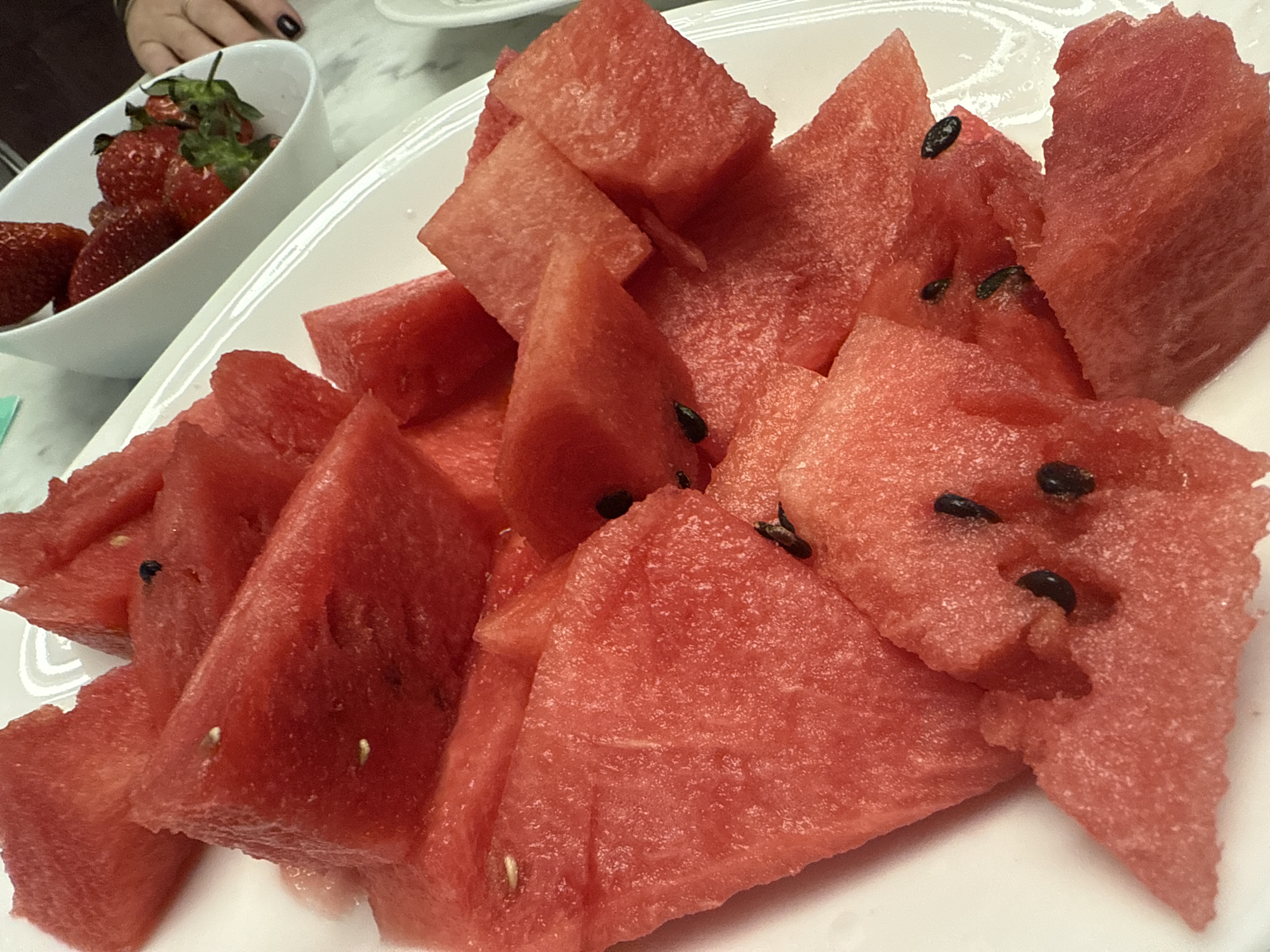A white plate holds several pieces of watermelon, cut into cubes and triangles, some containing black seeds. A white bowl containing strawberries is visible in the background. The scene suggests a meal or snack setting indoors. It is not possible to determine the location as Saint Petersburg, Russia, from the image alone. There is no information regarding people, actions, or interactions.