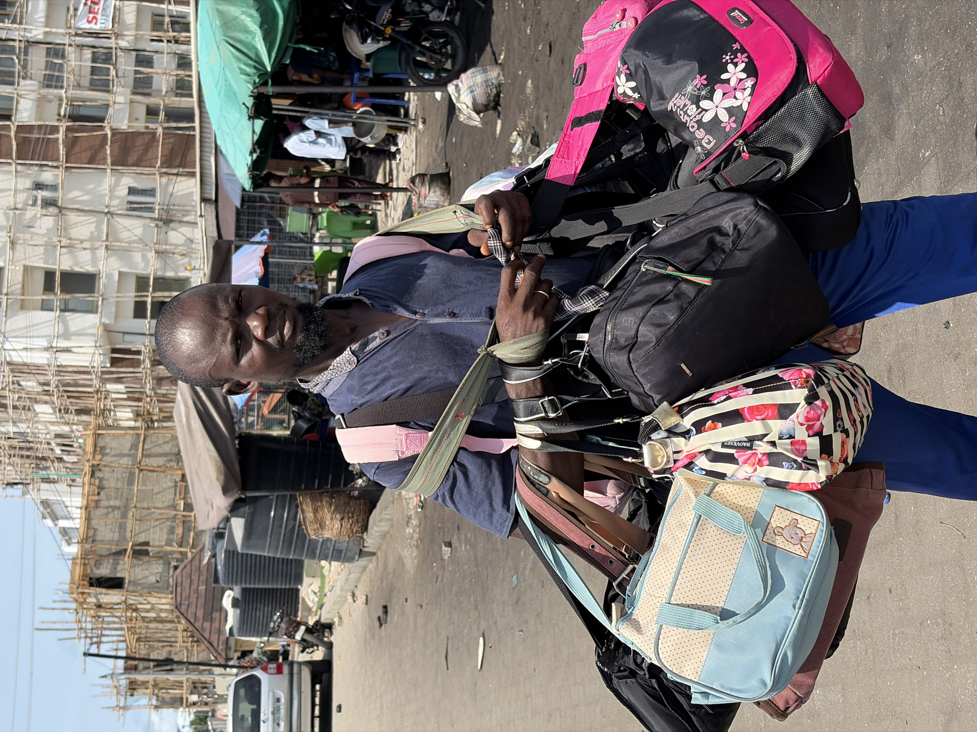 The image depicts a man in Igboefon, Nigeria, carrying a large stack of bags. He is wearing a navy blue shirt and blue pants. The bags vary in color and design, including pink, black, floral, and a light blue bag with a teddy bear applique. He is standing in a street, with buildings and scaffolding visible in the background. The street appears to be a busy area, with a vehicle, motorcycles, and people present.