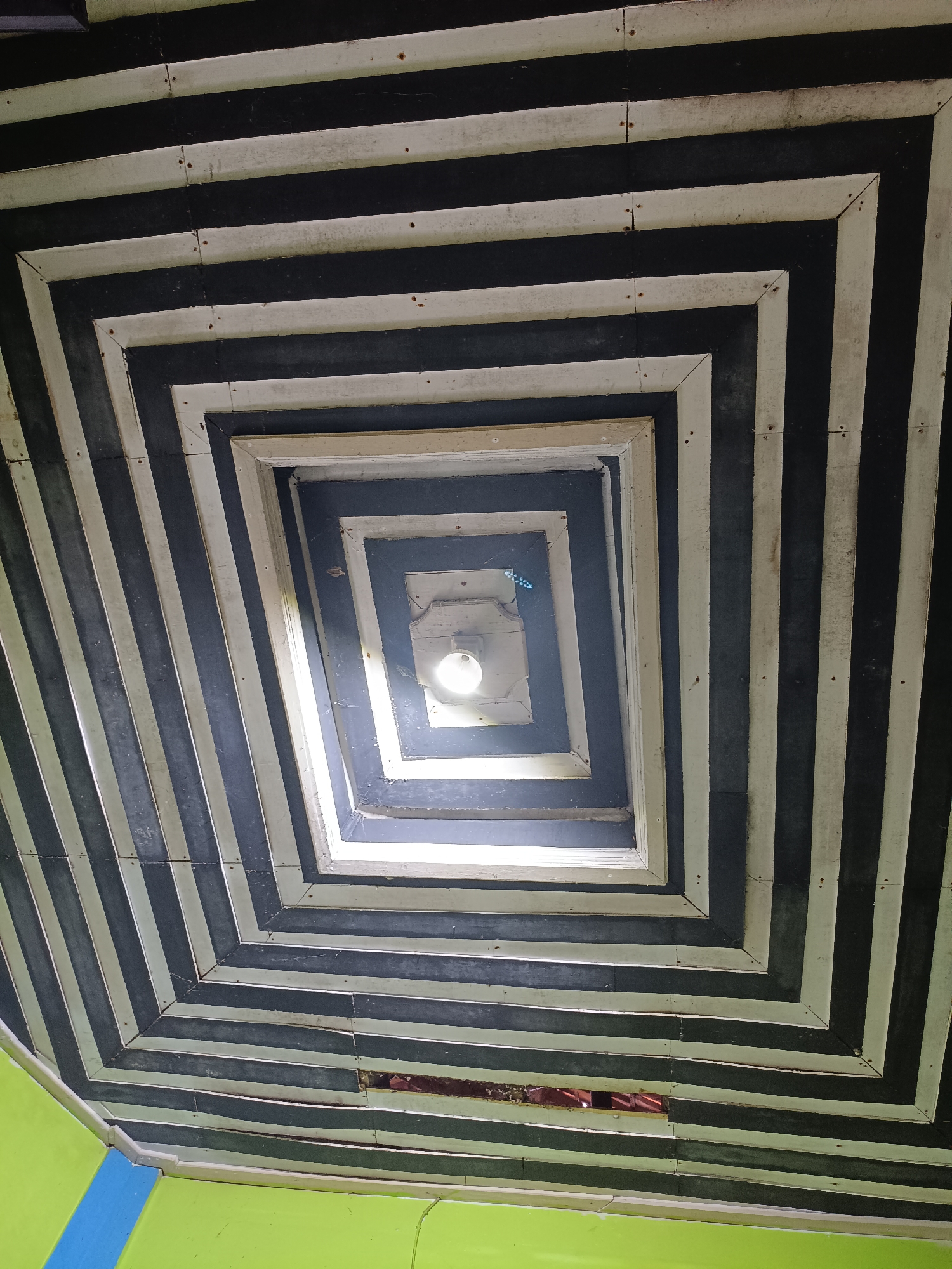 The image shows a view looking up at a geometric ceiling structure with alternating black and white rectangular panels. The structure appears to be an interior ceiling with a light fixture in the center. The walls below are painted green and blue. The location is identified as Kadununggal, Indonesia.