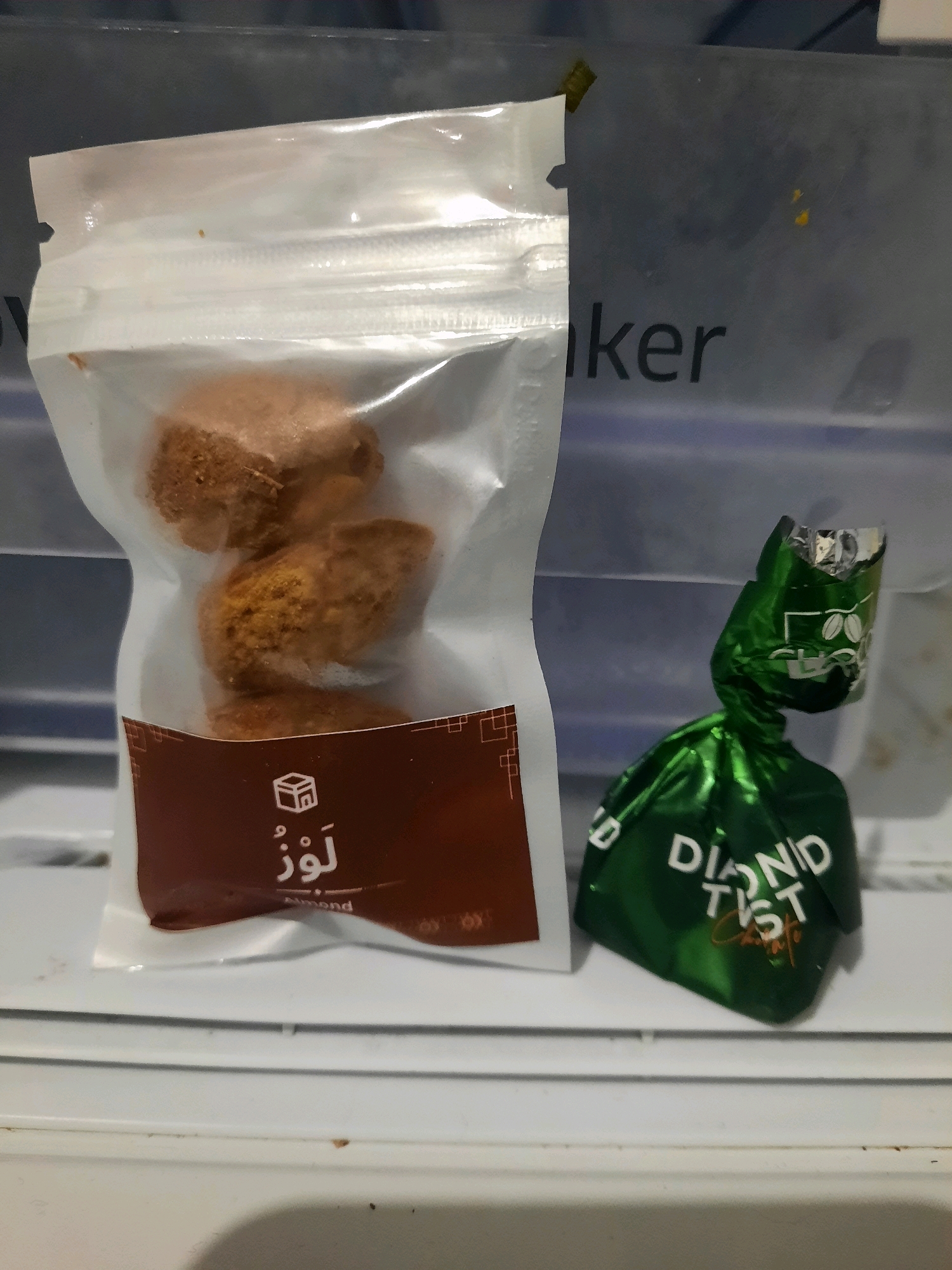 The image shows two food items on a white surface, possibly a shelf. On the left, there is a clear plastic bag containing several oval-shaped, brown, baked goods. The bag has a resealable top and a brown label at the bottom with white text and a small icon. On the right, there is a green, individually wrapped candy with white text that appears to say "DIAMOND TASTE". The background is a dark, textured surface with some light reflecting on it. The overall setting suggests an indoor environment, likely a kitchen or pantry. There is no indication of the city of East Jakarta, Indonesia in the image.