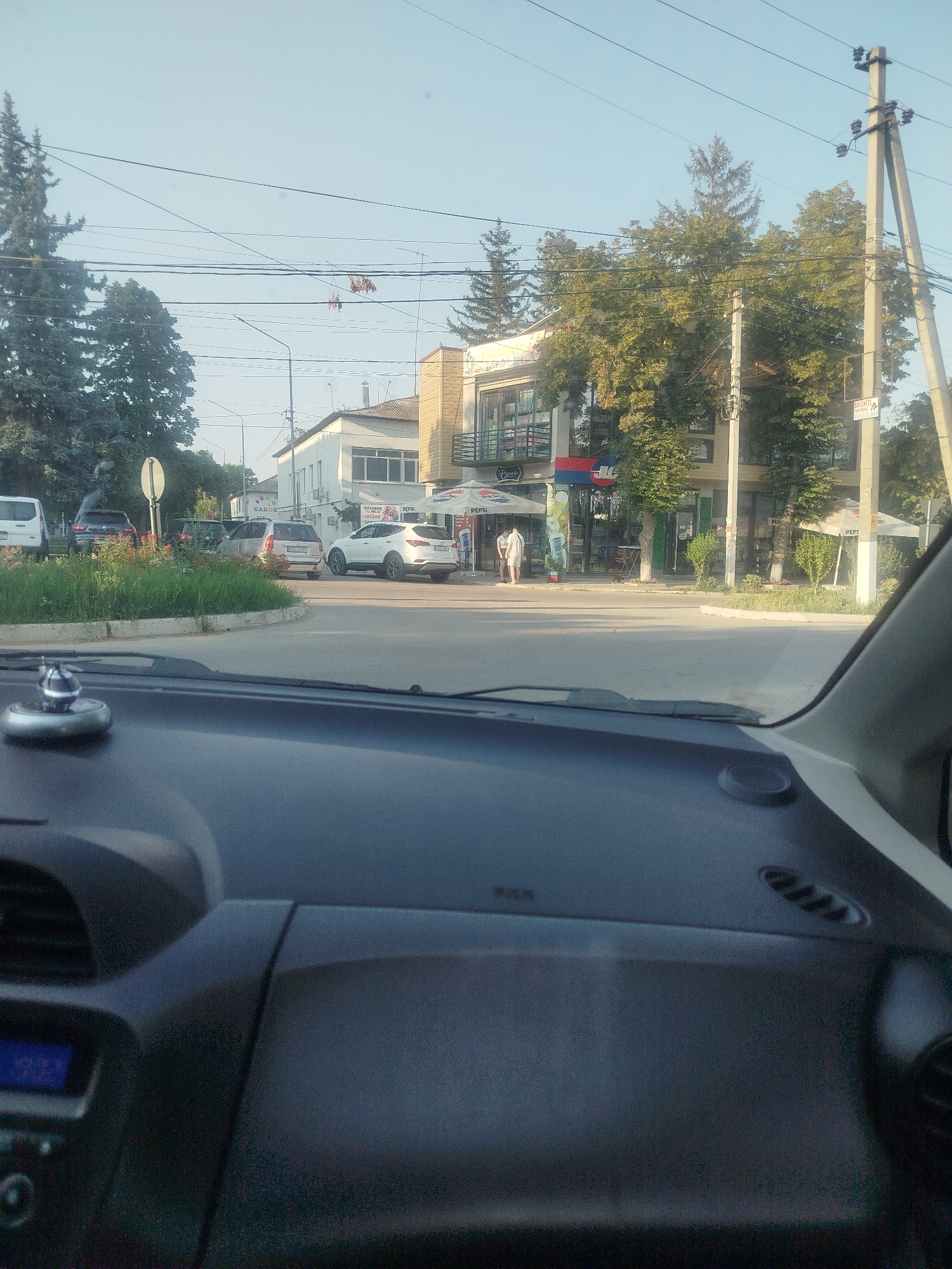 This media file depicts an outdoor scene viewed from inside a vehicle, likely in Criuleni, Moldova. The sun is shining, suggesting it is daytime, possibly late afternoon or early evening, given the soft light. The sky is clear and blue, indicating fair weather.
The scene is a street intersection with several buildings and vehicles. In the foreground, the dashboard of a car is visible, including a digital display showing "9.31". A small, decorative silver object is perched on the dashboard.
Further into the scene, there are several cars parked and in motion. These include a white minivan, a dark SUV, and a silver hatchback. A white SUV is also turning at the intersection. A circular flower bed with red and green foliage is prominent in the middle of the intersection.
Buildings line the street. One building appears to be a commercial establishment with a modern facade, featuring glass windows and balconies, and signage that includes the word "PEPSI" under a blue umbrella. Another building is a white, two-story structure with visible windows and air conditioning units. There are also numerous trees with green foliage, some quite tall and mature, interspersed throughout the landscape and surrounding the buildings. Power lines crisscross the sky, supported by concrete utility poles.
Two people are visible walking outside one of the buildings. They appear to be casually dressed. One is wearing a light-colored shirt and shorts, and the other is wearing a white outfit.
A sign on a utility pole has some visible text, including what appears to be "BATTE" followed by numbers, and another sign in Cyrillic script. The overall impression is a typical street scene in a town or city.