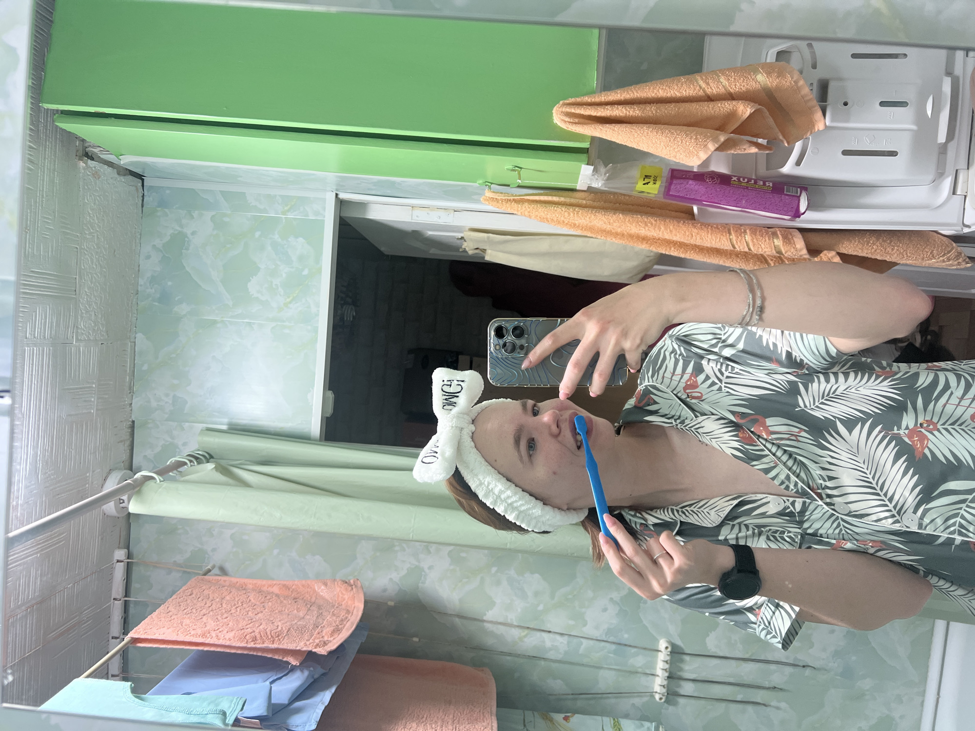 The image depicts a young adult female taking a mirror selfie in a bathroom located in the city of Chaikovsky, Russia. The woman, with light skin and light brown hair pulled back by a white terrycloth headband with a bow and "OWO" text, is wearing a short-sleeved pajama top patterned with green palm leaves and red flamingos. She is holding a blue manual toothbrush to her mouth, appearing to brush her teeth. Her left wrist displays a black smartwatch and a small white mark on her forearm. A silver ring is visible on her right ring finger. She holds a silver-colored smartphone with a clear, subtly patterned case and three visible camera lenses in her right hand, aimed at the mirror.
The bathroom environment, reflected in the mirror, features light green walls, possibly clad in patterned PVC panels or tiles with a subtle, swirling texture. The ceiling consists of white, textured panels. On the left side of the reflection, a white extendable clothes drying rack holds a light blue towel and a light orange towel. A white circular object, likely a smoke detector or ceiling fixture, is visible further right on the ceiling. A light green shower curtain hangs from a rod in the mid-ground. On the right, a bright green wall-mounted cabinet with two doors, one slightly ajar, is prominent. Two light orange towels hang from gold-colored hooks attached to this cabinet. A purple bottle with Cyrillic text ("БЕТОХ") is suspended in a clear plastic bag from another hook, adjacent to the towels. Below these items, the white top-loading dispenser tray of a washing machine is visible. In the background, reflected through an open doorway, a dimly lit area with exposed brick-like walls and a white door frame is discernible, with a cream-colored tote bag hanging on a hook.