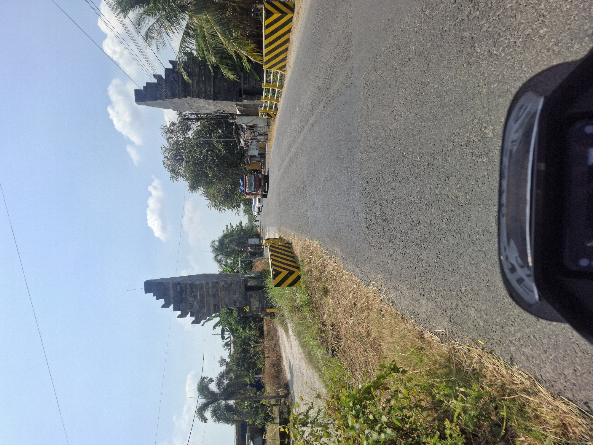 The image depicts a road leading through a gate-like structure in Karangcangkring, Indonesia. On either side of the road are tall, stone structures resembling gateways with stepped tops. Yellow and black striped barriers are placed along the side of the road. Palm trees and other vegetation are visible. A red truck and several cars are on the road, and two motorcycles are parked to the right. The sky is blue with scattered clouds. The perspective suggests the photo was taken from a moving vehicle.