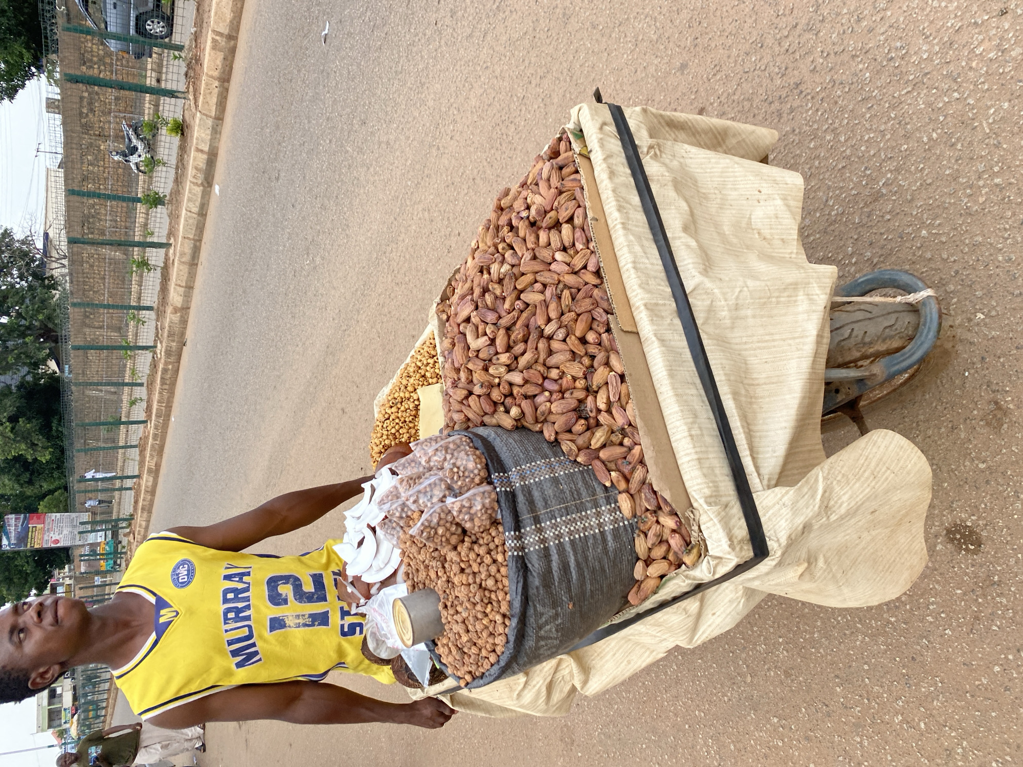 In Bukuru, Nigeria, a young Black man wearing a yellow basketball jersey with "MURRAY" and the number "12" displayed prominently is pushing a cart laden with various food items. The cart is covered with a beige cloth, draped over a frame. The man's jersey also has a "DVC" logo. The cart's contents include a large black bag overflowing with small, round, brownish nuts. Several smaller plastic bags filled with similar nuts are placed on top of the larger bag. To the right of the black bag are piles of dried dates, some in cardboard containers. In the background, a paved road runs alongside a sidewalk bordered by a green fence and a stone wall. Beyond the wall, trees and buildings are visible. A white motorcycle is parked along the roadside. Other pedestrians and a vehicle are visible in the distance. The ground is a mix of asphalt and dirt.