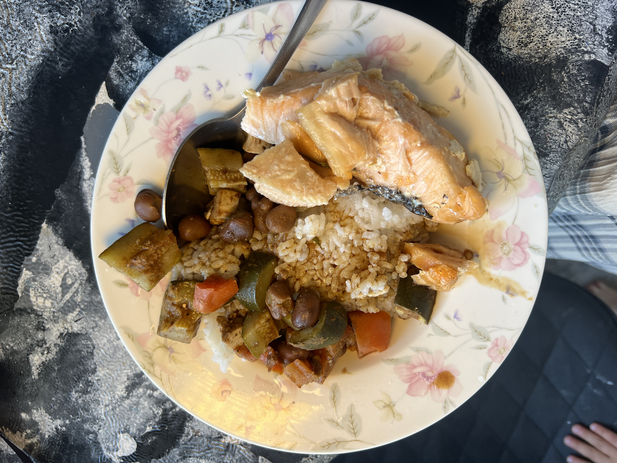 An overhead shot displays a meal served on a white ceramic plate adorned with a floral pattern of pink and purple flowers and green leaves. The plate contains cooked salmon, partially flaked and light orange-pink, with some skin visible on one piece. Alongside the salmon is white rice, parts of which are mixed with a brownish sauce or seasoning. A medley of cooked vegetables and beans accompanies the rice, including diced green vegetables (likely zucchini or cucumber), diced orange carrots, and dark brown beans, all integrated into a savory sauce. A silver-colored metal spoon rests on the left side of the plate, partially submerged in the food. The meal is situated on a table surface featuring an abstract, textured design predominantly in black, white, and grey, suggestive of a resin or epoxy art finish. The scene is illuminated by natural daylight, indicating a daytime setting. A small portion of a hand is faintly visible in the bottom right of one of the crops. There is no visible text in the image.
