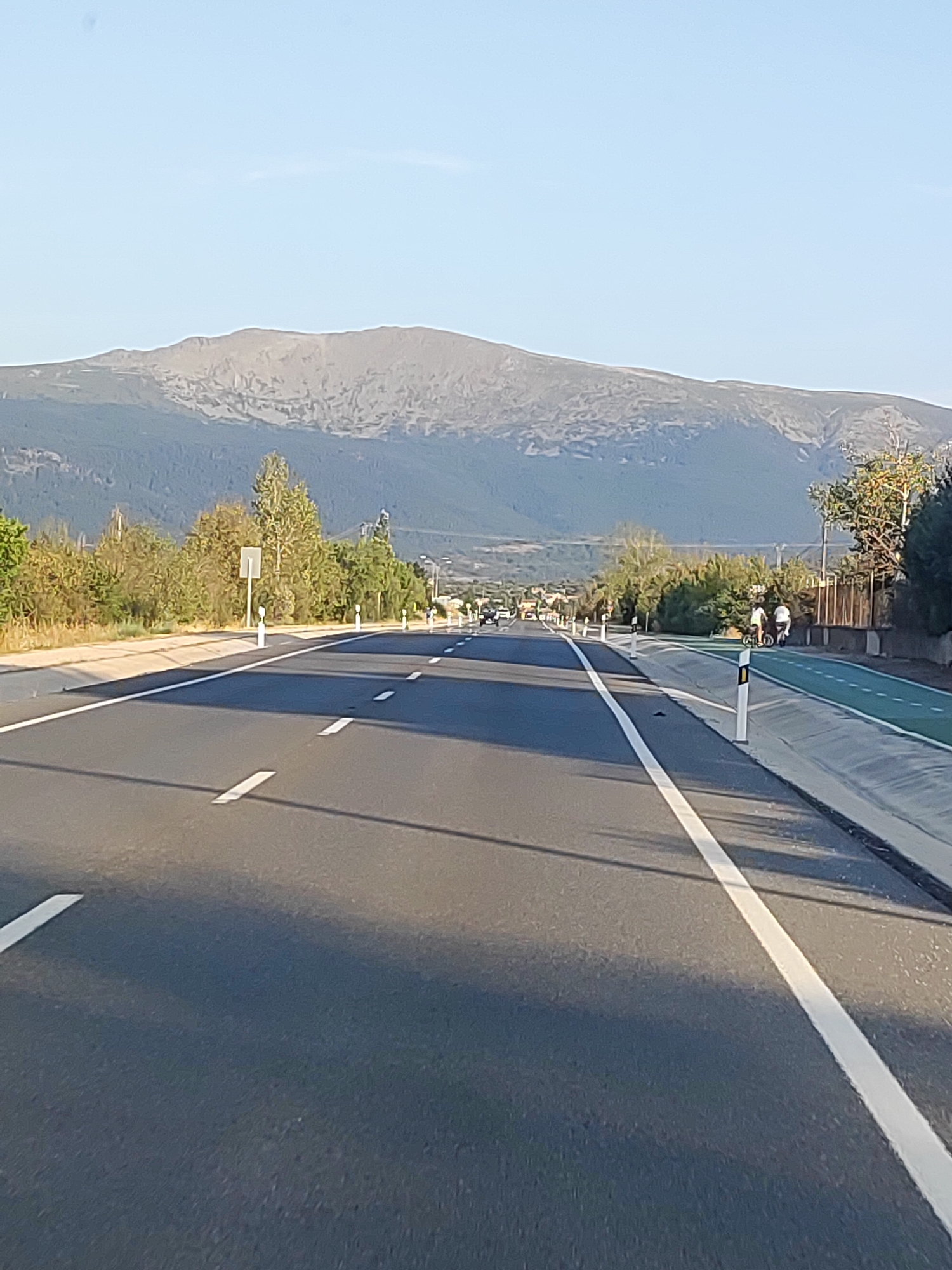 A view down a multi-lane highway towards a distant mountain range under a clear blue sky. The road is asphalt with white lane markers. To the right of the highway is a green-colored bicycle lane separated by a concrete barrier and bollards. Two individuals are visible on the bicycle lane, one riding a scooter-like device and another walking. Vegetation lines the sides of the highway and the bicycle lane. In the far distance, a large, rugged mountain range dominates the horizon. The image appears to be taken from a moving vehicle, as evidenced by the dynamic perspective and the shadow of the vehicle cast on the road. The location is identified as Carrascalejo, Spain.