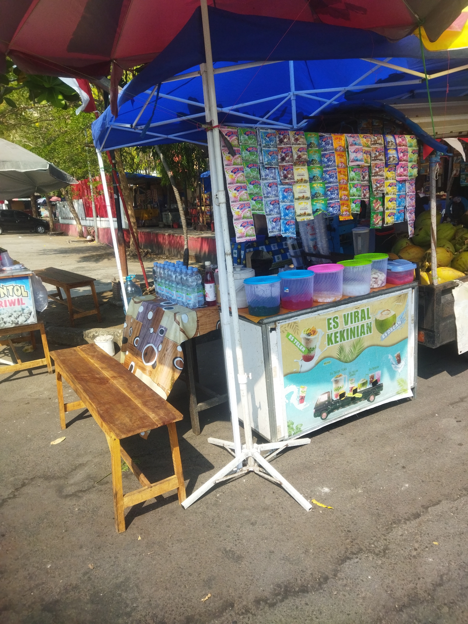 An outdoor refreshment stand is set up under a canopy in what appears to be the city of Wonogiri, Indonesia. The stand features a variety of drinks, including bottled water and colorful beverages in clear plastic containers. Packets of instant drink mixes are displayed hanging from the canopy, with labels in Indonesian. In the foreground, wooden benches are placed for customers. Behind the stand, a truck with a load of green fruits, likely coconuts, is visible. The ground is paved and shows signs of being a public area, possibly a roadside or a market. The lighting suggests it is daytime, with sunlight casting shadows.