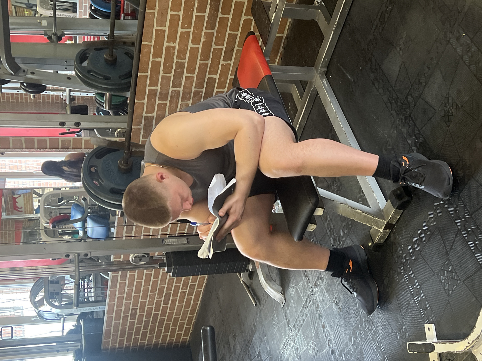 A young adult male sits on a weight bench in a gym, holding and writing in a notepad with a pen. He is wearing a gray tank top, black Kappa shorts, and black athletic shoes with orange accents. The gym has brick walls and black rubber flooring. Weightlifting equipment is visible in the background. There is no indication of the city or country.