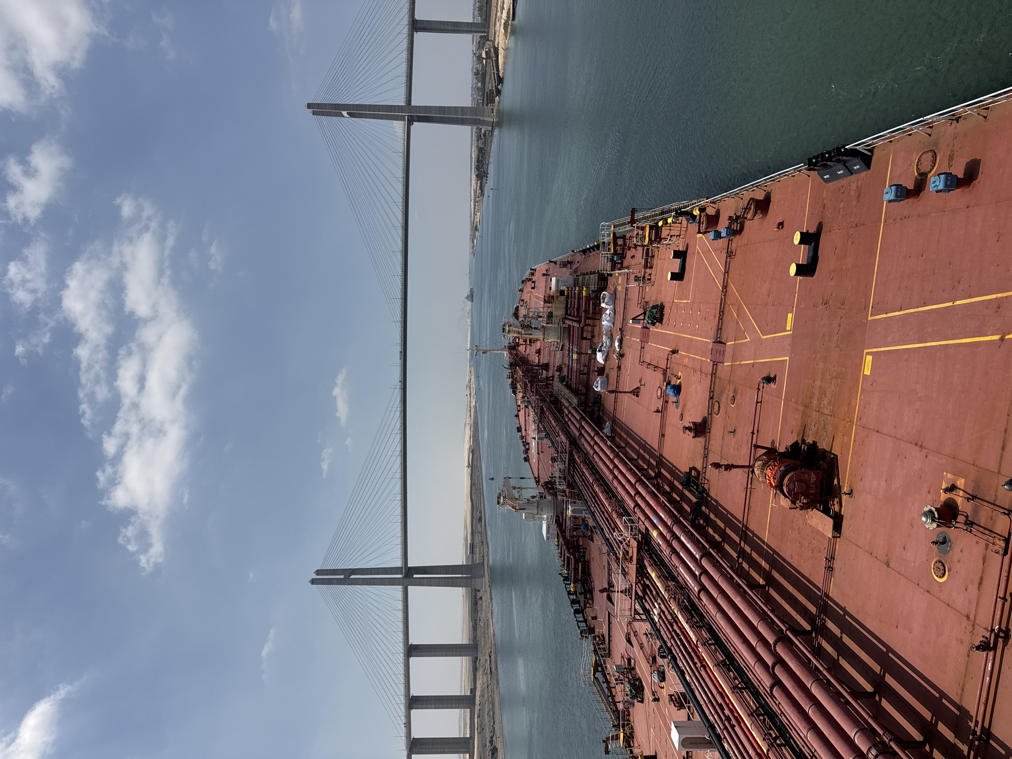 The image captures a perspective from the deck of a large red cargo ship, likely a tanker, as it navigates a wide, calm body of water. Ahead, a massive cable-stayed bridge spans the water, connecting two banks of sandy terrain. The bridge features two tall, V-shaped pylons supporting numerous cables that hold up the deck. The water is a deep blue-green, reflecting the bright, partially cloudy sky. On the ship's deck, a complex network of red pipes and various pieces of industrial equipment are visible. In the distance, other vessels can be seen, and the banks of the waterway are lined with sparse vegetation and structures. The scene appears to be the Suez Canal, specifically in the vicinity of Qantara West, Egypt, given the presence of the large bridge and the characteristic landscape. There are no visible individuals interacting in the frame.