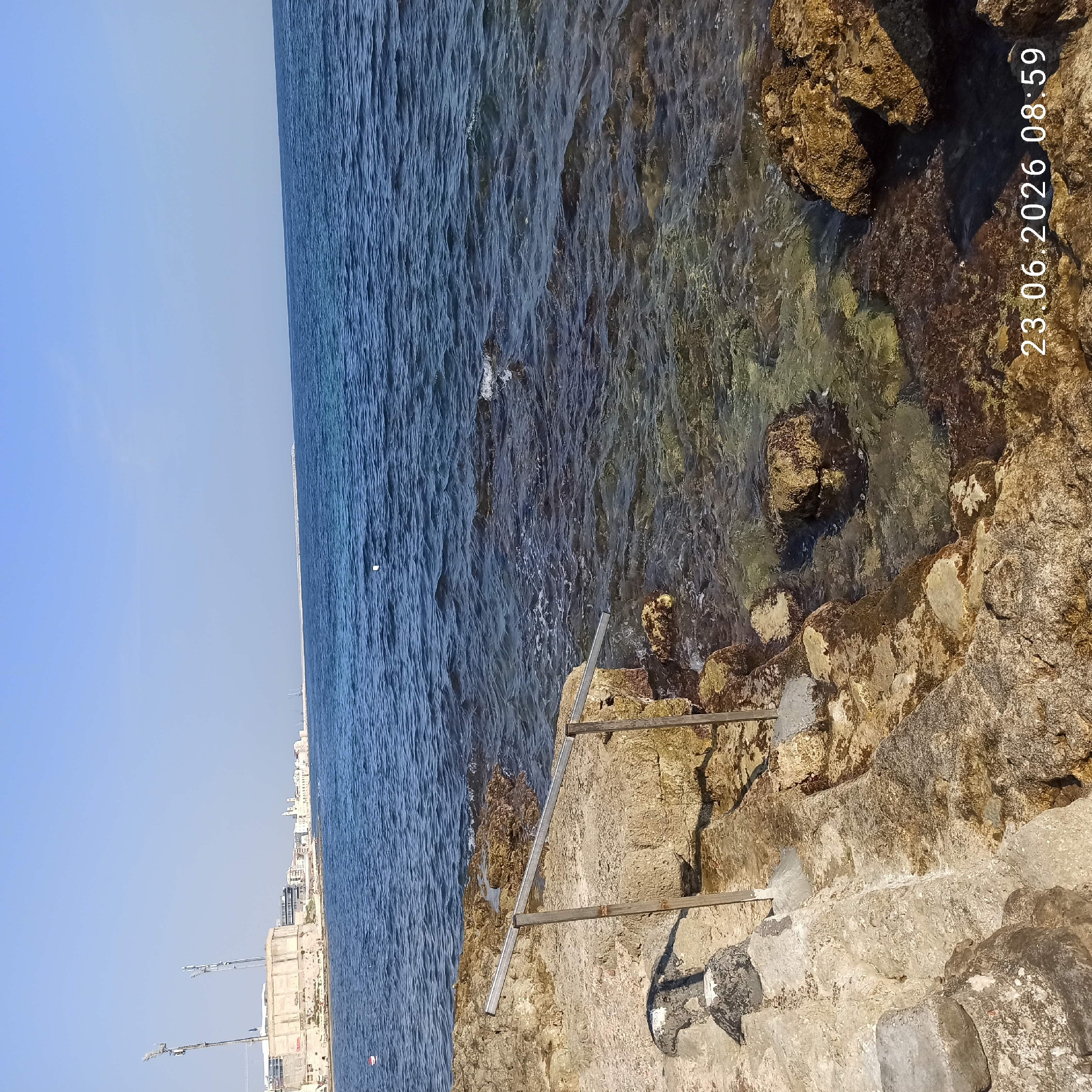 The image captures a view of the Molfetta coastline in Italy, featuring a clear blue sky and calm sea. In the foreground, rugged, light-colored rocks form the coastline, with some areas submerged by the transparent water. A metal railing with two upright supports is visible, offering a pathway or support structure along the rocky edge. The water is shallow near the shore, allowing the seabed and rocks beneath the surface to be seen. In the distance, a cityscape stretches along the horizon, characterized by a line of buildings and a prominent, modern-looking structure with two tall antennas. The presence of a white floating object in the water suggests a marker or buoy. The timestamp "23.06.2026 08:59" is overlaid on the bottom right of the image.