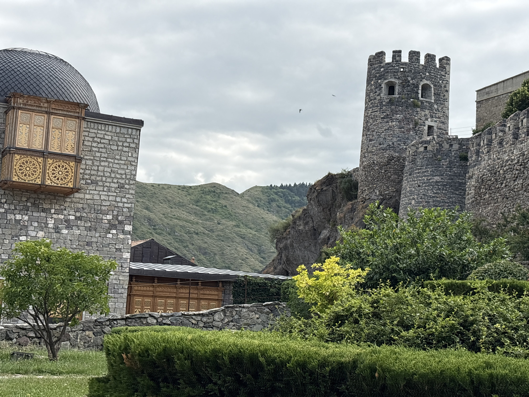 The image shows the Rabati Castle complex in Akhaltsikhe, Georgia. The scene is dominated by historical architecture and natural landscape under an overcast sky.
On the left side of the frame, a section of a stone building is visible, topped by a dark, patterned dome. Attached to this building is a wooden balcony or oriel window, elaborately carved with geometric patterns in shades of gold and brown. The stone walls of the building are constructed from rectangular blocks.
To the right, a round, stone watchtower with crenellated battlements and arched windows stands prominently. Adjacent to it is a section of a fortified stone wall, also made of rough-hewn stones.
In the foreground, a manicured garden features various green shrubs and bushes, with a small, leafy tree to the left. A low stone wall runs through the garden. Behind the architecture and garden, rolling green hills rise, covered in dense vegetation and trees. The sky is cloudy, with two birds visible in flight. A wooden building with decorative carvings is partially visible behind the stone wall and tree. The overall impression is one of a historic site set within a lush, mountainous environment.