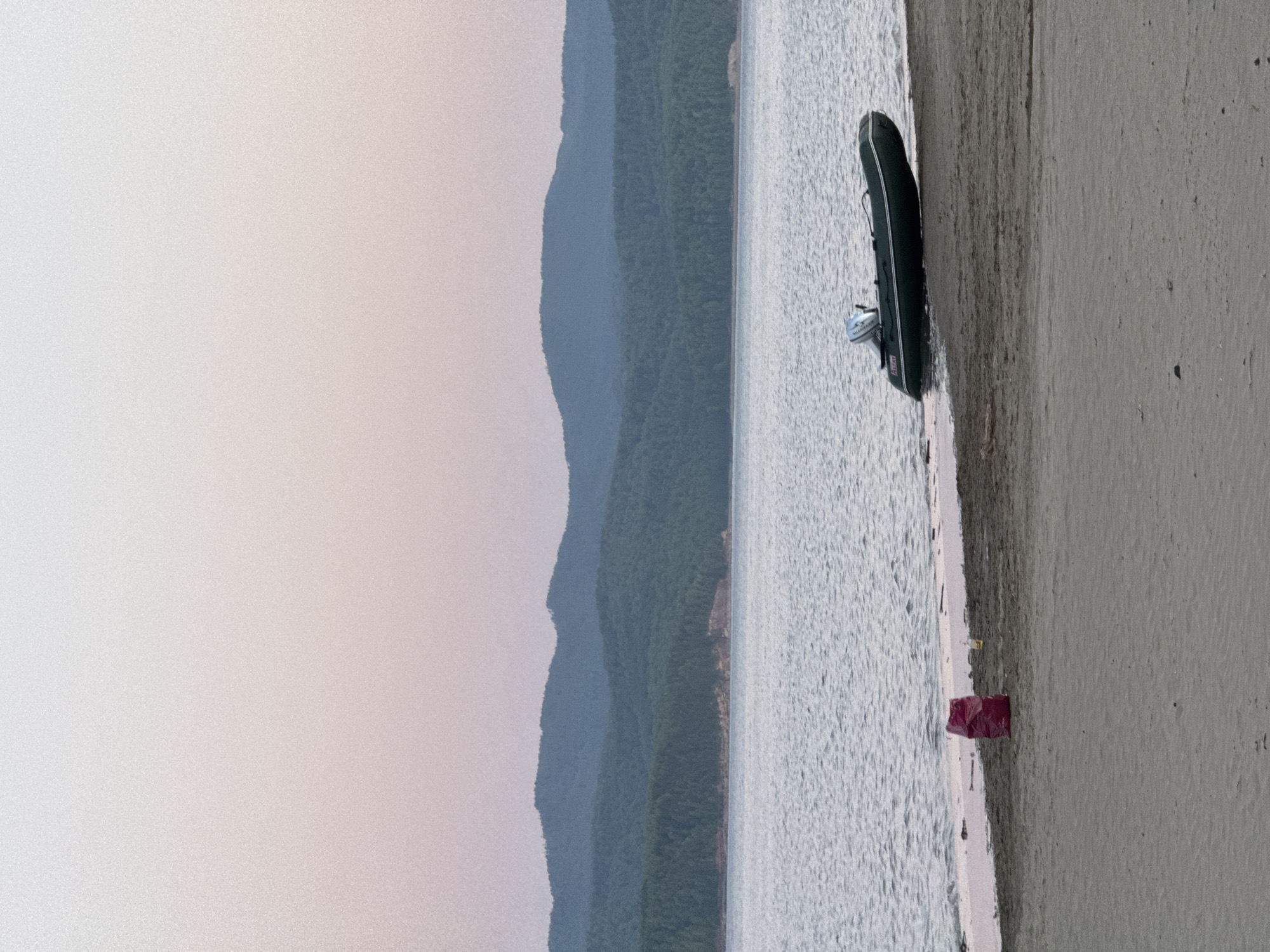 The image depicts a serene lakeside or reservoir scene at Daurskoe, Russia, captured during either dawn or dusk. The sky above presents a gentle gradient, transitioning from a light, almost white tone at the zenith to a soft pinkish-purple hue near the horizon. Below the sky, a series of forested hills and mountains, appearing dark blue-green, stretch across the mid-ground, their silhouettes distinct against the pastel sky.
The expansive body of water, likely a lake or reservoir, occupies the middle section of the frame, exhibiting a rippled surface with a grayish-blue reflection of the ambient light. The water meets a sandy shore in the foreground, characterized by light brown to grayish-tan sand.
On the right side of the sandy shore, a dark green inflatable boat with white accent stripes is beached, partially resting in the water and partially on the sand. A light-colored outboard motor is visibly attached to the stern of the boat, raised out of the water. On the left side of the shore, closer to the water's edge, a red plastic bag, possibly containing refuse, stands upright. Slightly behind and to the right of the red bag, a clear or light-colored bottle lies on the sand. No individuals are present in the scene, which conveys a quiet and unoccupied atmosphere.