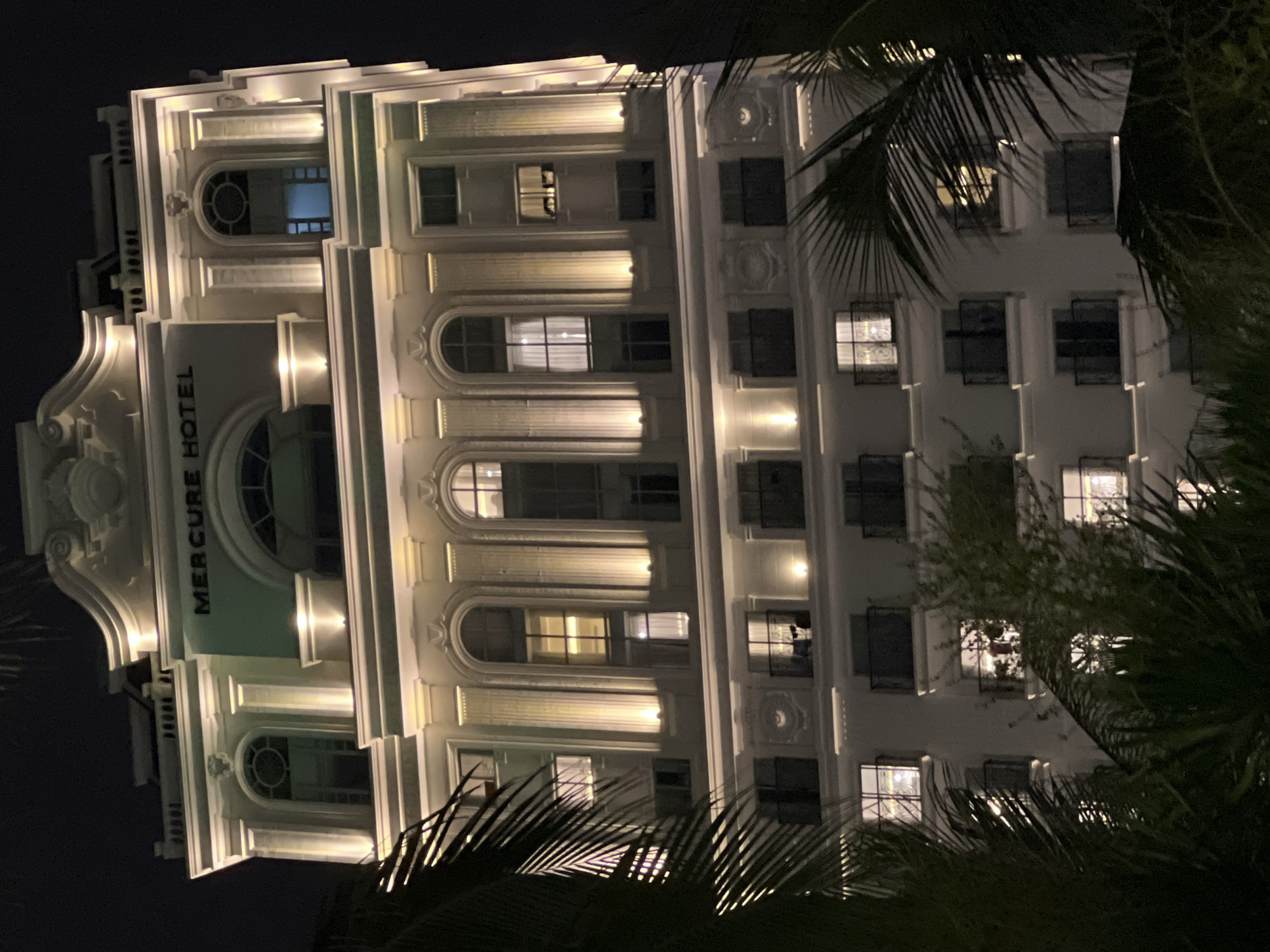 The image shows the Mercure Hotel in Nha Trang, Vietnam, at night. The hotel is a tall, white building with ornate architectural details and is illuminated by warm yellow lights. Palm fronds are visible in the foreground, obscuring parts of the lower floors of the hotel. Several windows on the hotel are lit, suggesting occupancy. The sky is dark, indicating it is nighttime.