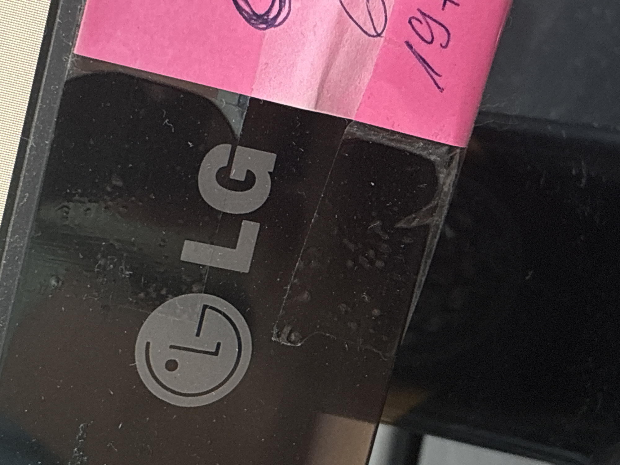 The image shows a close-up view of a black electronic device, likely a monitor, with the LG logo visible on its surface. The LG logo consists of a stylized circular icon representing a face and the letters "LG" next to it. A bright pink sticky note with handwritten markings in purple marker is attached to the right side of the device, partially covering the "G" of the LG logo. The sticky note has what appears to be a date written on it, possibly "19+". The device surface appears to be dusty and has some smudges. The background is dark, and the top of the image shows a sliver of a textured, light-colored surface, possibly the screen of the device or another object. The scene is a close-up shot, with no wider environmental context provided.