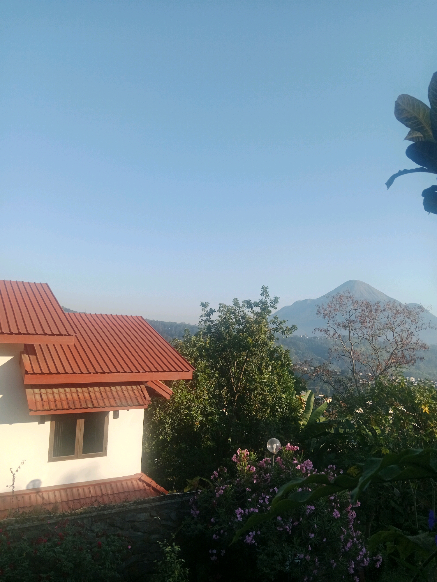 This image captures a serene landscape scene with a modern house in the foreground, set against a backdrop of lush greenery and a majestic mountain. The house features a prominent red-tiled roof and white walls, with a window visible. Surrounding the house is a garden filled with flowering plants and trees. In the distance, a conical mountain dominates the horizon, shrouded in a light haze. The sky is a clear, bright blue. The scene appears to be located in or around Prigen, Indonesia, given the architectural style, vegetation, and the presence of a mountain typical of the region. The overall atmosphere is peaceful and natural, suggesting a quiet morning or late afternoon.