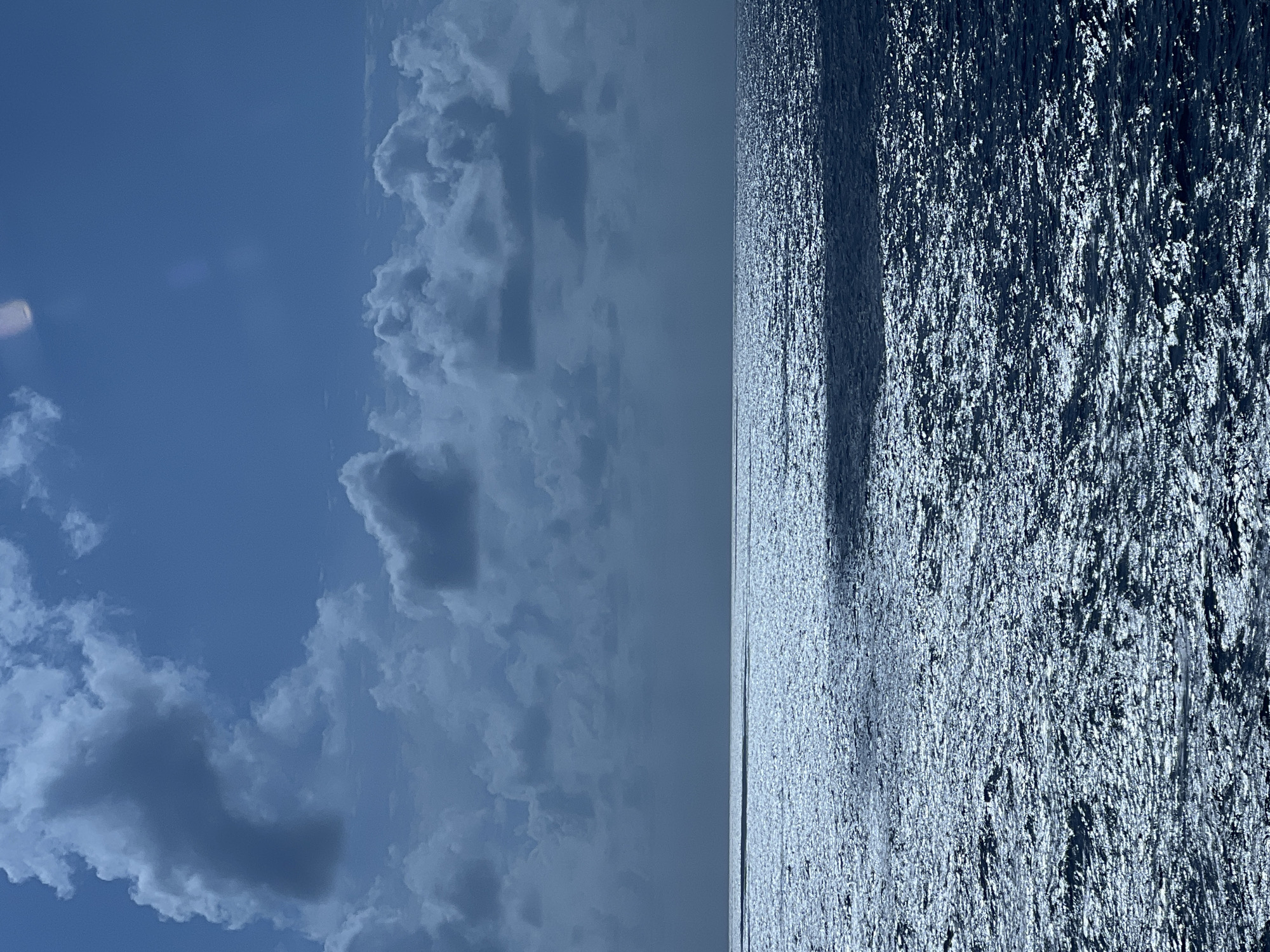 A wide-angle shot captures a vast expanse of the ocean under a partially cloudy sky. The sea is active, with sunlight glinting off the choppy surface, creating a shimmering effect. The horizon is a sharp line dividing the deep blue water from the sky, which is filled with dramatic, dark clouds and patches of brighter sky. The overall impression is one of a dynamic and somewhat moody seascape. The image does not provide any information about a specific city or location.
