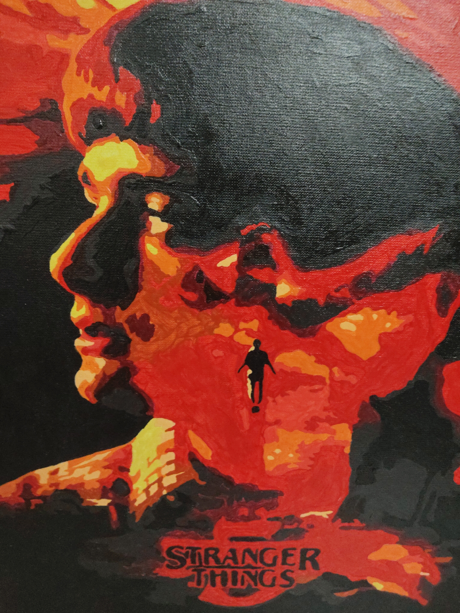 The image is a stylized painting on canvas, featuring a large profile of a human head, predominantly in black, facing left. The interior of the head is filled with an abstract, vibrant landscape of intense reds, oranges, and yellows, resembling a fiery or otherworldly terrain. Visible within this red-orange landscape, a small, silhouetted figure of a person appears to be walking or running across the rugged ground. The figure is rendered entirely in black, contrasting sharply with the warm tones around it.
The painting style exhibits visible brushstrokes and the textured weave of the canvas, suggesting an acrylic or oil medium. The colors are stark and high-contrast, with deep black serving as both the background and the main outline of the head, against the vivid, almost glowing interior. The overall impression is one of mystery, tension, and a journey through a fantastical or dangerous environment.
At the bottom center of the artwork, the text "STRANGER THINGS" is clearly visible in a distinctive red, stylized font. This text identifies the media as related to the popular television series, implying the scene depicted within the head is evocative of the show's "Upside Down" dimension or similar supernatural elements. The time of day and weather are not explicitly depicted due to the abstract nature of the artwork, but the color palette suggests a dramatic, non-natural setting.