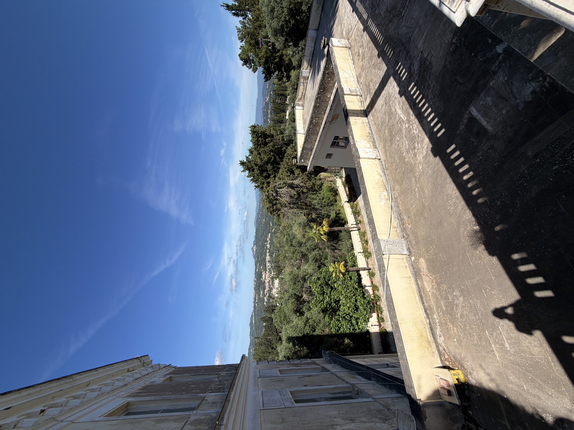 The image presents a panoramic view from a elevated position, looking out over a verdant landscape under a bright blue sky. The scene is bathed in clear, strong daylight, casting distinct, elongated shadows, suggesting it is either morning or late afternoon. The location is Gastouri, Greece.
In the foreground and to the right, a concrete or paved terrace is visible, extending away from the viewer. A white railing runs along its edge, casting prominent, striped shadows across the textured surface of the terrace. A shadow that appears to be cast by the photographer is also visible in the bottom right corner. A small yellow utility box is attached to a wall in this area.
To the left, a large, ornate building with multiple windows and decorative architectural details rises. Its weathered appearance suggests age. Further down the slope in the middle ground, another smaller, light-colored building is nestled amidst dense green foliage. The landscape beyond is characterized by rolling hills thickly covered in trees and shrubs, including some palm trees. In the distance, a valley or coastal plain unfolds, dotted with numerous small structures that form a town or settlement. The horizon is marked by distant hills, and the sky above is a clear, vibrant blue with scattered wispy white clouds.
No explicit activity is taking place; the scene is static, showcasing the architecture and natural beauty of the region. There is no visible legible text.