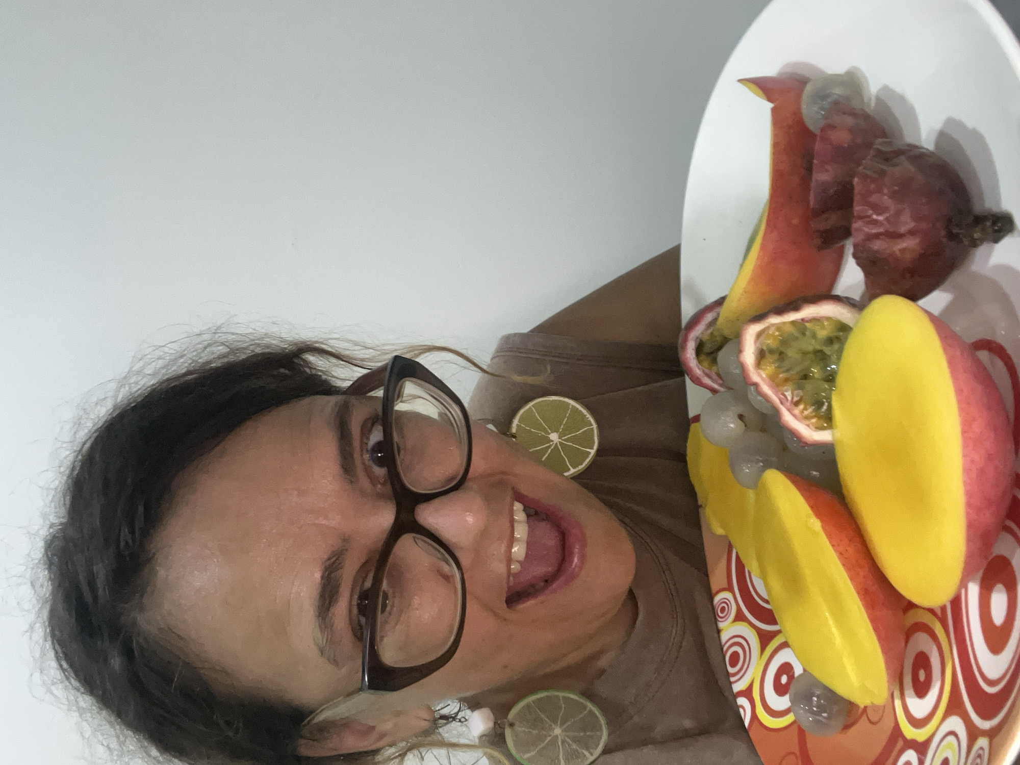 A woman with dark, curly hair and dark-rimmed glasses looks directly at the camera with her mouth open in a surprised expression. She is wearing a light brown t-shirt and has lime slice earrings. In front of her is a white plate filled with various fruits, including sliced mangoes, passionfruit, grapes, and what appear to be dried plum halves. The plate is positioned on a colorful, circular patterned surface. The background is a plain, off-white wall. The location is not specified in the image or context provided.