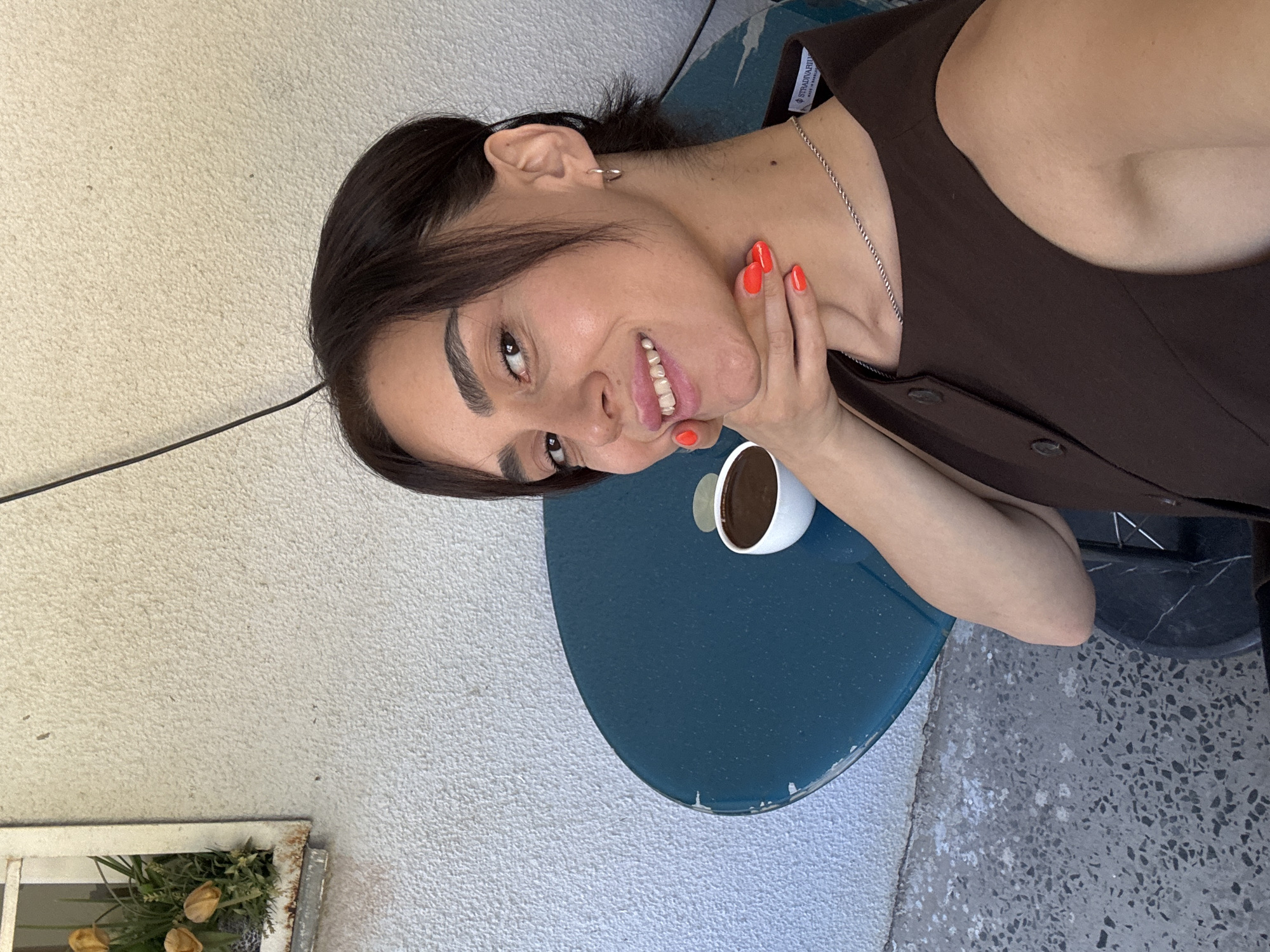 In an outdoor setting, a woman with dark hair and a fair complexion takes a selfie. She is wearing a brown sleeveless top with buttons and a silver chain necklace. Her nails are painted a vibrant orange. Her right hand is resting on her chin, and she is smiling. In front of her, on a blue round table, is a small white cup filled with a dark liquid, likely coffee. To the left of the woman and behind her, a section of a textured white wall is visible, with a small window box containing artificial flowers and greenery. The ground appears to be a grey, speckled surface. The overall impression is a candid moment captured during a casual outdoor break, possibly in Tbilisi, Georgia, given the architectural and environmental cues.