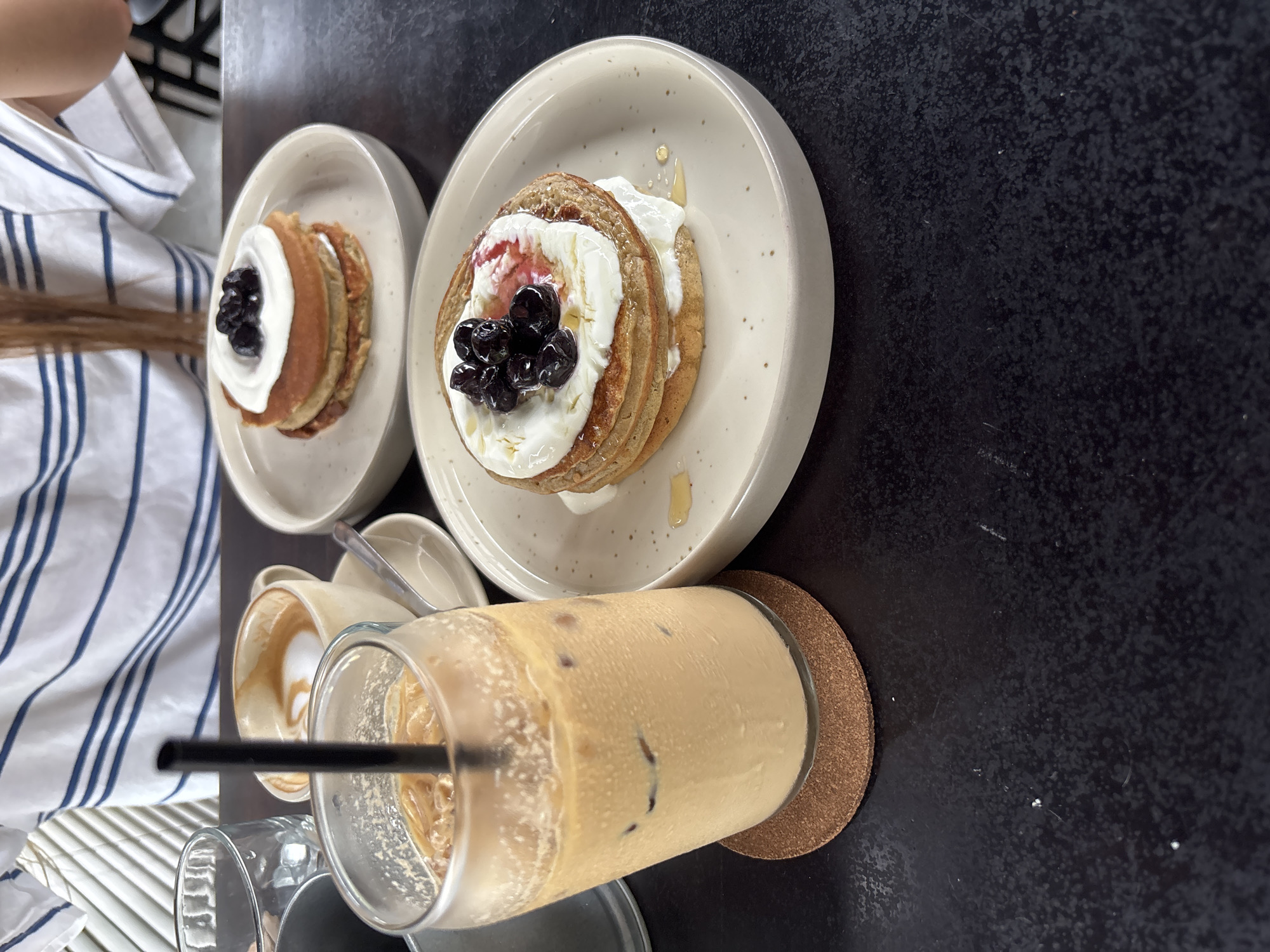 The image presents an overhead view of a cafe table set with breakfast or brunch items in Kuta Selatan, Indonesia. The dark, textured tabletop holds two plates of pancakes, an iced coffee, and other beverage containers.
Two plates, off-white with speckled patterns, each feature a stack of three small pancakes. The pancakes are generously topped with a white cream or yogurt and garnished with dark blueberries. One stack also shows a hint of reddish fruit topping beneath the blueberries. Both plates display drizzles of a golden liquid, likely honey or syrup, around the edges.
In the foreground, a clear glass of iced coffee, light brown in color with ice cubes, stands on a round, cork coaster. A black straw is inserted into the drink. Behind it, a ceramic cup with visible latte art suggests another coffee drink. An empty glass is also partially visible on the left.
A person is seated at the table, with their upper torso, left arm, and long, light brown hair visible on the left side of the frame. They are wearing a white shirt with vertical blue stripes. The lighting suggests it is daytime. No discernible text is visible on any objects. The scene conveys a relaxed meal or snack in a casual setting.