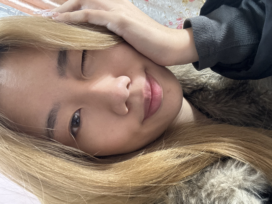 A close-up, matter-of-fact portrait of a single individual is depicted in an indeterminate indoor location within Shapingba District, China. The subject is a young female with long, straight, light blonde or light brown hair that frames her face. She is facing directly towards the viewer. Her right eye is closed in a wink, while her left eye is open. A slight smile is present on her lips. Her left hand is raised, with the palm resting against her left cheek and jawline. The fingers of her left hand are visible, and her fingernails are elongated with a speckled, light-colored design. She is dressed in an outer garment with visible light brown faux fur trim, worn over a dark grey, ribbed long-sleeved shirt. The background is out of focus, showing a light-colored surface with subtle, faint floral or patterned details visible on the right side.