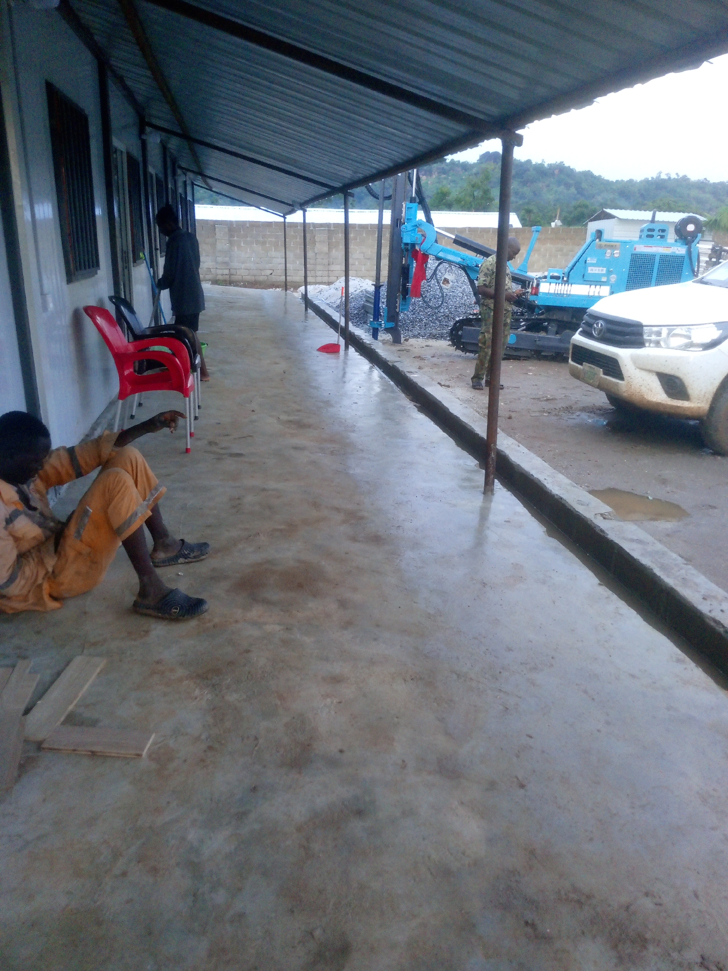 The image depicts an outdoor scene under a covered walkway, likely at a construction or work site, with a concrete floor that appears wet. In the foreground to the left, a person wearing an orange jumpsuit with reflective stripes is sitting on the ground, facing away from the camera and pointing with their right hand. Behind this person, three chairs are visible, two red and one black. Further back, a person in a grey jacket is standing and appears to be sweeping or cleaning. In the background, a blue drilling rig is parked next to a white pickup truck. The area is surrounded by a low concrete block wall and lush green hills can be seen in the distance. The overall impression is of a functional, possibly rural, setting in Indo, Nigeria.