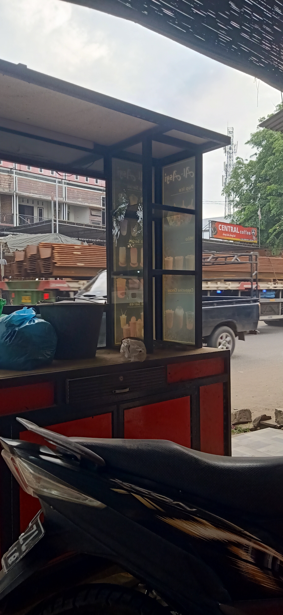 The image captures a street-level view in Gampongteungoh, Indonesia, focusing on a small roadside kiosk selling drinks. The kiosk has glass panels displaying images of beverages and has a red and black base structure. In the foreground, the seat and rear of a dark-colored motorcycle are visible, suggesting a common mode of transport. Behind the kiosk, a truck with a load of what appear to be metal pipes is driving on the road, and beyond that, buildings and trees form the urban backdrop. A prominent red sign with white lettering, "CENTRAL coffee," is visible on a pole in the distance. The sky is overcast, indicating possibly cloudy weather.