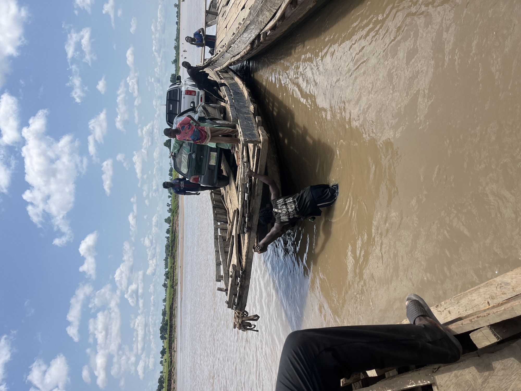 An outdoor, medium shot captures a ferry operation on a wide, muddy river under a cloudy sky in Nemnei, Nigeria. Two wooden barges, tied together, are positioned side-by-side. A green sedan is loaded onto the left barge, while a white pickup truck is on the right barge. Several individuals are present. On the left barge, a person is partially submerged in the water, reaching towards the edge. Another person, seen from the waist down, stands on the wooden planks of the left barge. On the right barge, two people are standing near the pickup truck, and another person is seated. In the distance, the riverbank is lined with green vegetation and trees. The water appears calm with ripples.