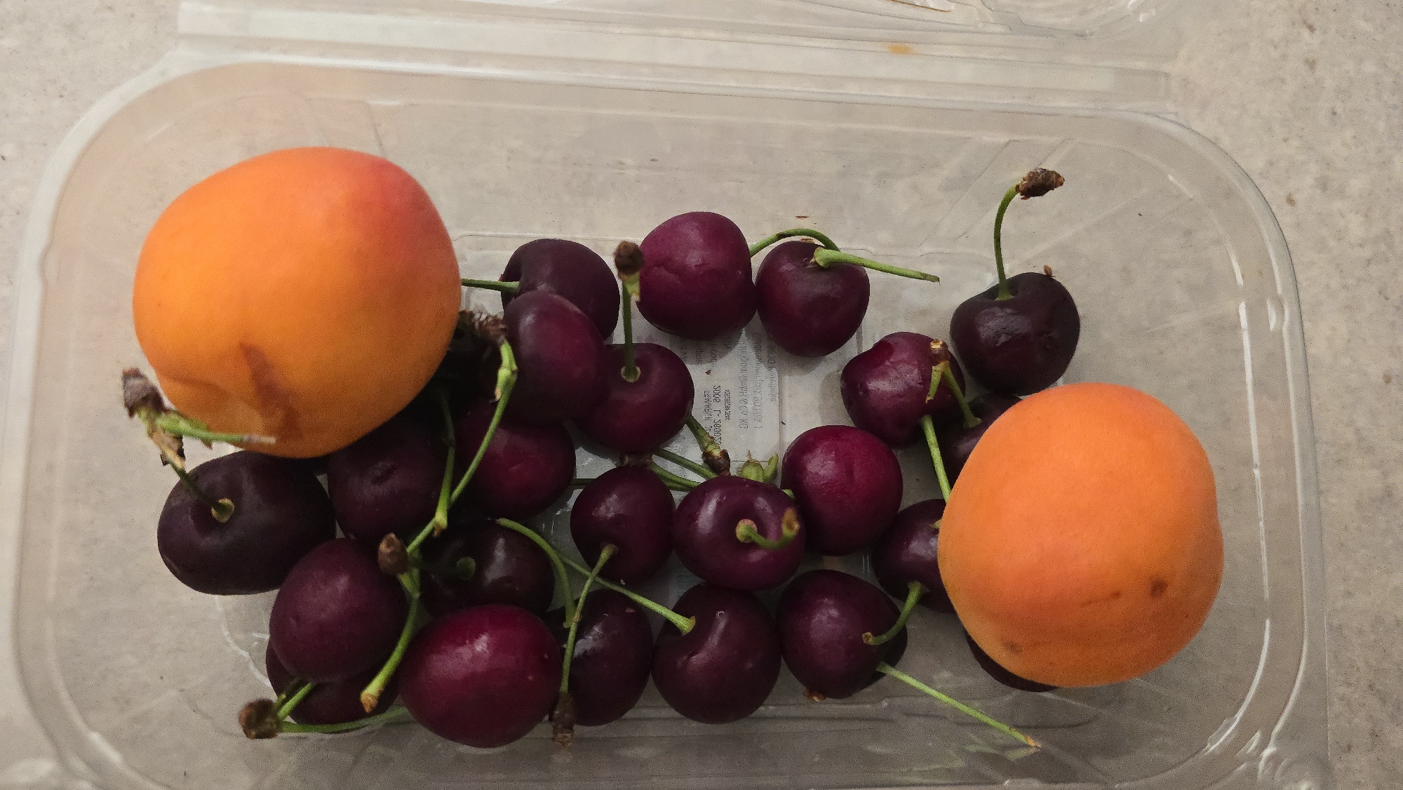 A clear plastic clamshell container holds a mix of dark red cherries and two orange apricots. The container sits on a light beige surface. Some of the cherries have their stems attached.