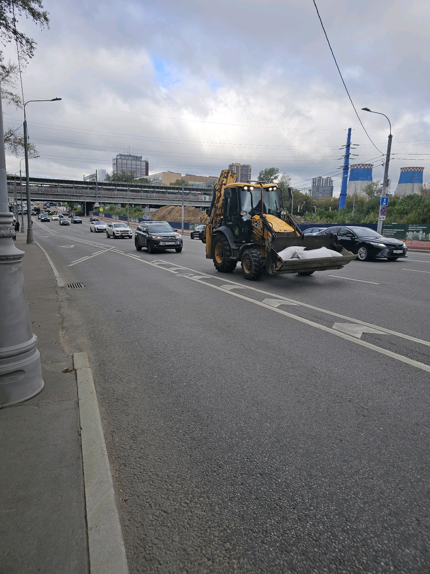 A JCB backhoe loader, carrying several white sandbags in its front loader, drives on a multi-lane asphalt road in Moscow, Russia. The road has white lane markings and appears to be an urban street or highway. Other vehicles, including cars and SUVs, are also present on the road. In the background, there is an elevated overpass, some buildings, power lines, and industrial structures, including cooling towers with blue and white checkered tops. The sky is overcast with scattered clouds. The scene suggests ongoing construction or road maintenance work.