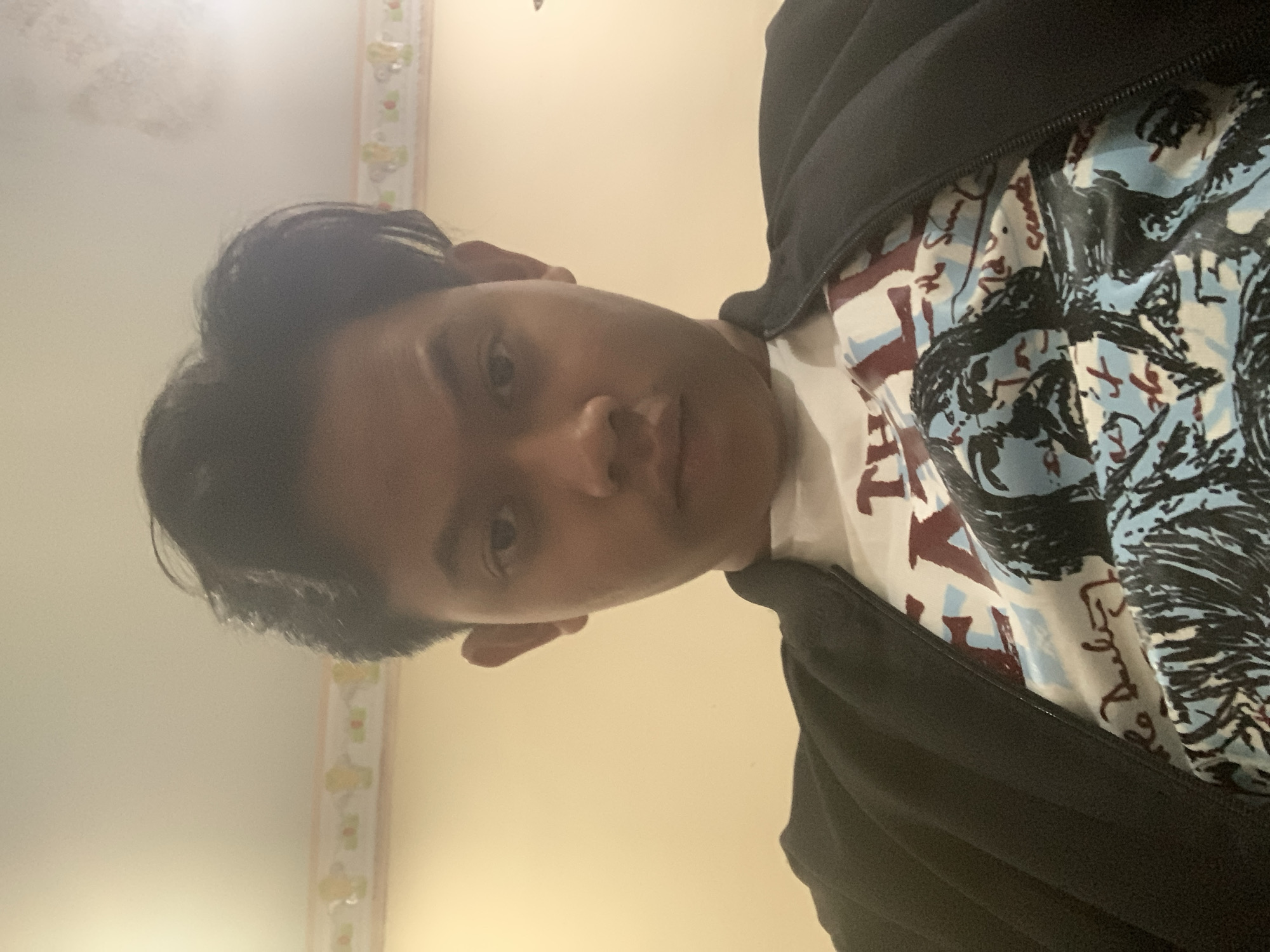 A young man, appearing to be of Southeast Asian descent, is captured in a low-angle, indoor selfie. He has dark, styled hair and a serious expression. He is wearing a black jacket over a white t-shirt adorned with a graphic print that includes abstract figures and text. The background is a plain, light-colored wall with a decorative border near the ceiling. The lighting suggests an artificial light source, likely from above. No specific landmarks or indications of the city of Dukuh Mojo, Indonesia, are visible within the frame.