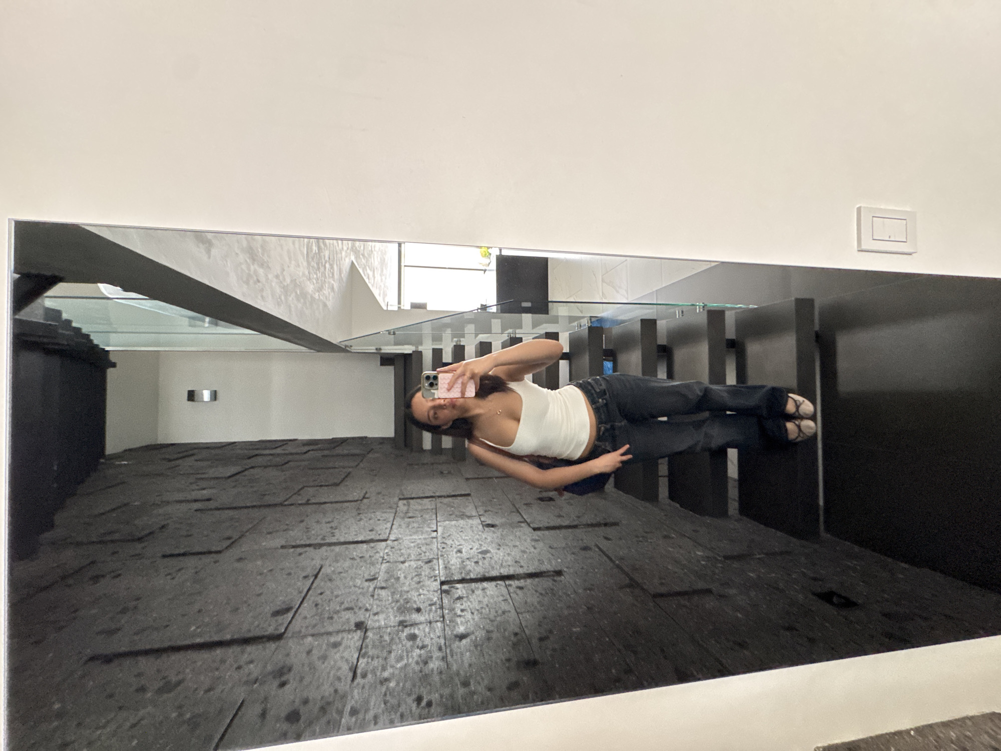 The image shows a reflection in a large mirror, likely attached to a wall in a building. The reflection features a young woman holding a mobile phone up to take a picture of herself. She is wearing a white tank top and dark jeans with sandals. The background reflected in the mirror includes dark tiled floors, glass partitions or railings, and some visible natural light. The building's interior appears modern. The image does not provide enough information to determine the precise location in Puebla, Mexico.